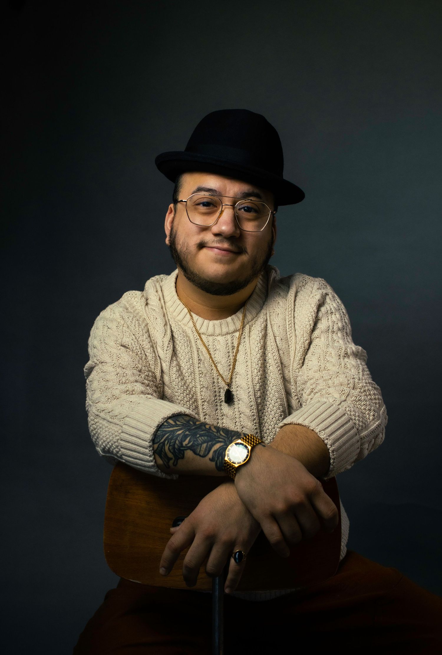 Portrait of someone in glasses and fedora wearing a cream sweater while resting on a wooden chair.