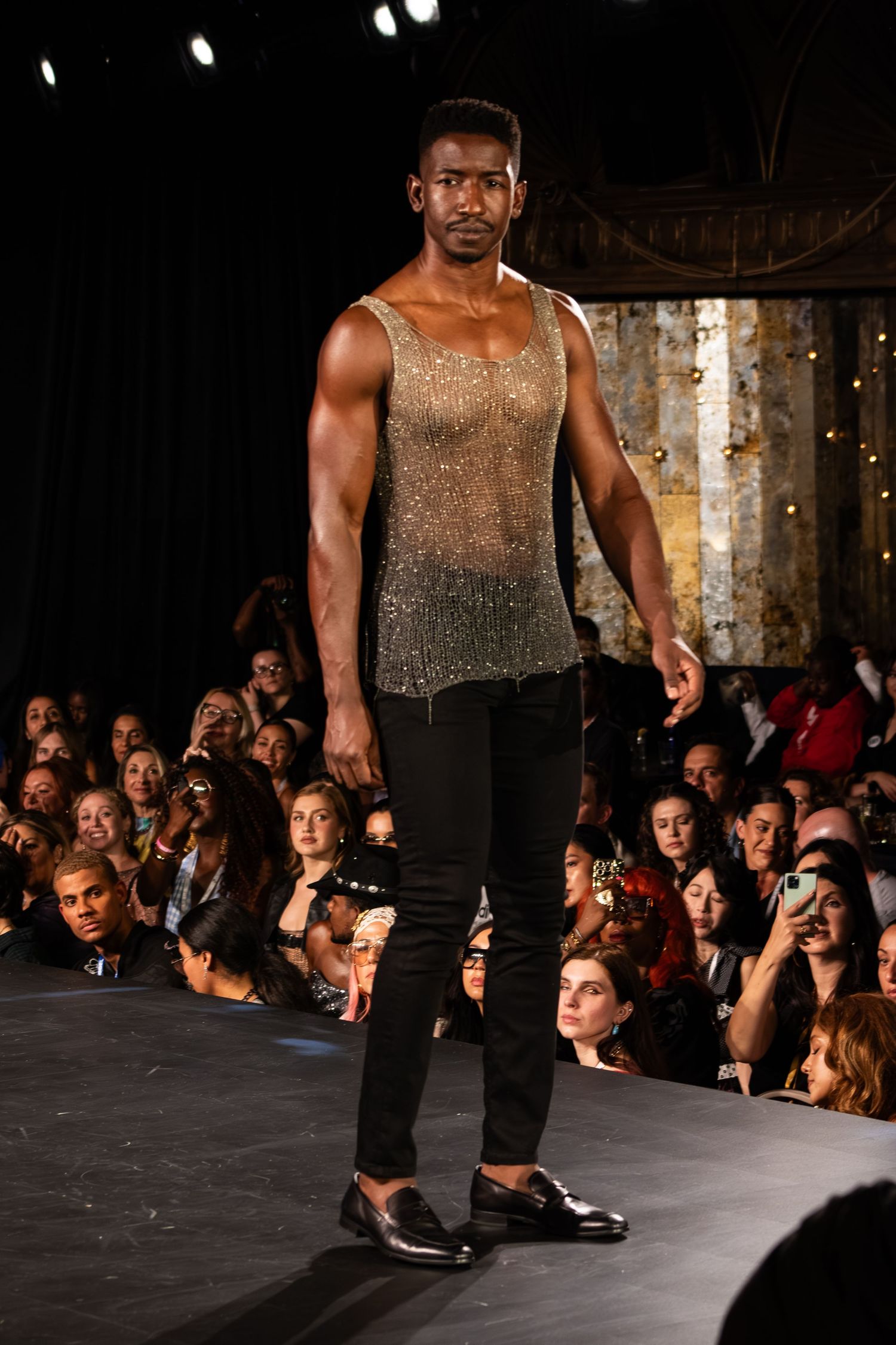 Muscular male model walks runway wearing sheer gold embellished tank top with black pants and loafers.