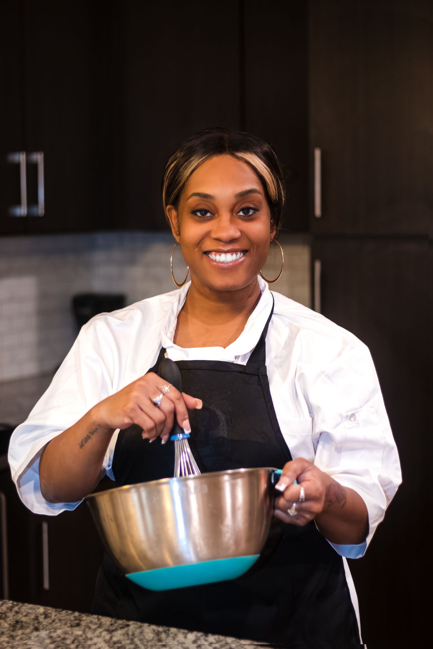 A chef in a black apron and white shirt whisks ingredients in a metal mixing bowl in a professional kitchen.