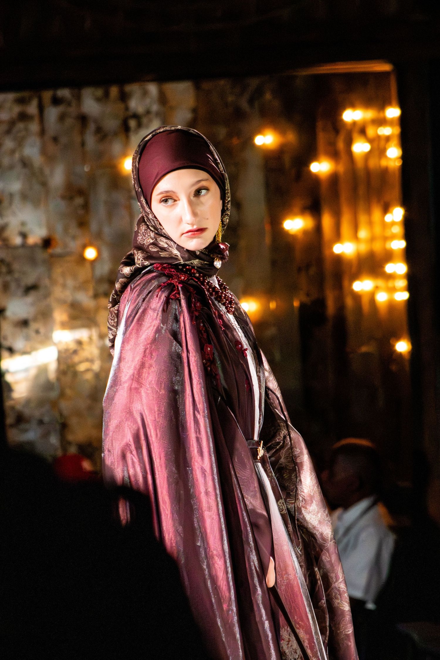 Model showcases burgundy velvet cape and headscarf on dark runway with ambient lighting.