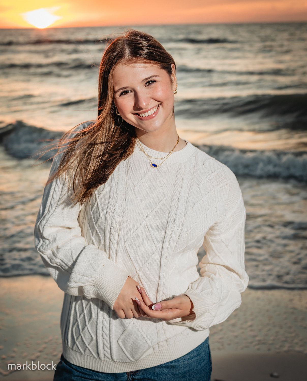 HS Senior girl in a white cable knit sweater stands smiling on a Naples FL beach at sunset as waves roll in behind them.
