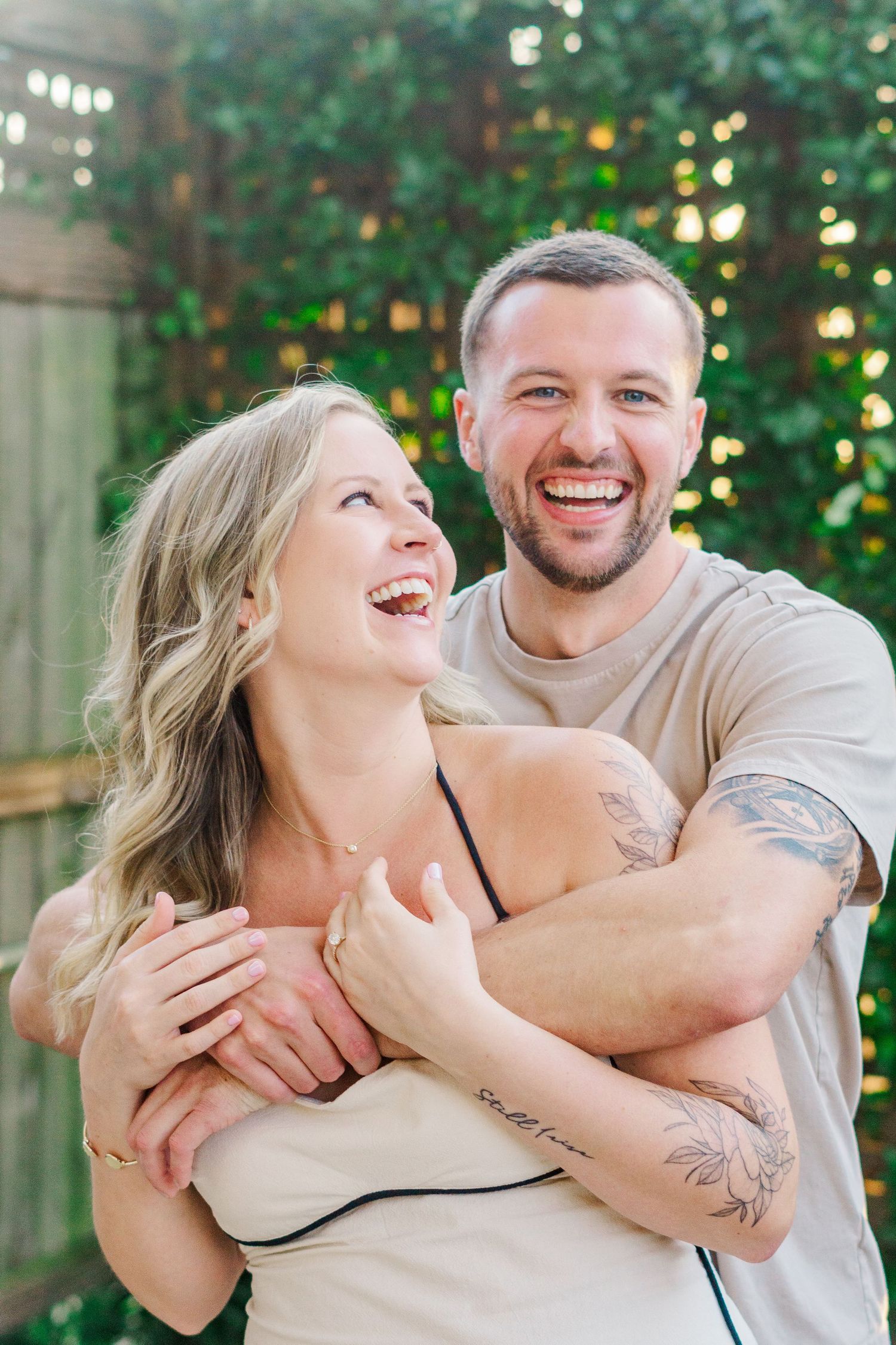 A joyful couple laughs and embraces in front of lush greenery and twinkling lights.