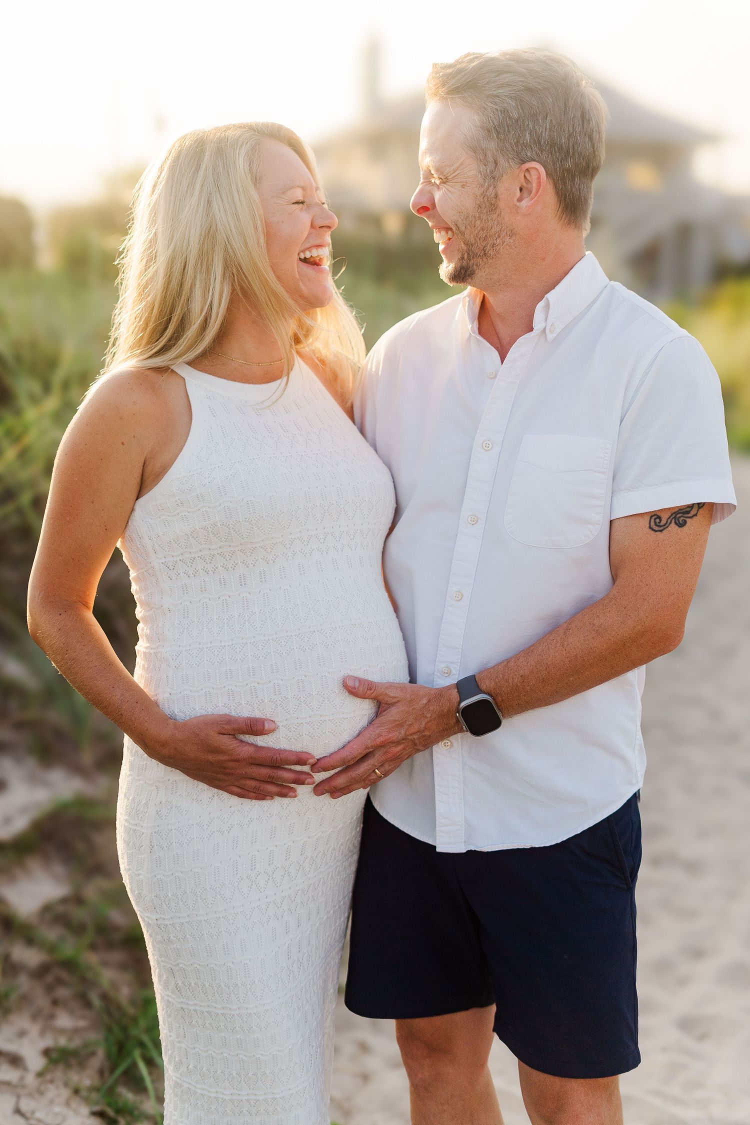 Joyful couple shares laughter during maternity photoshoot on beach path at dusk.