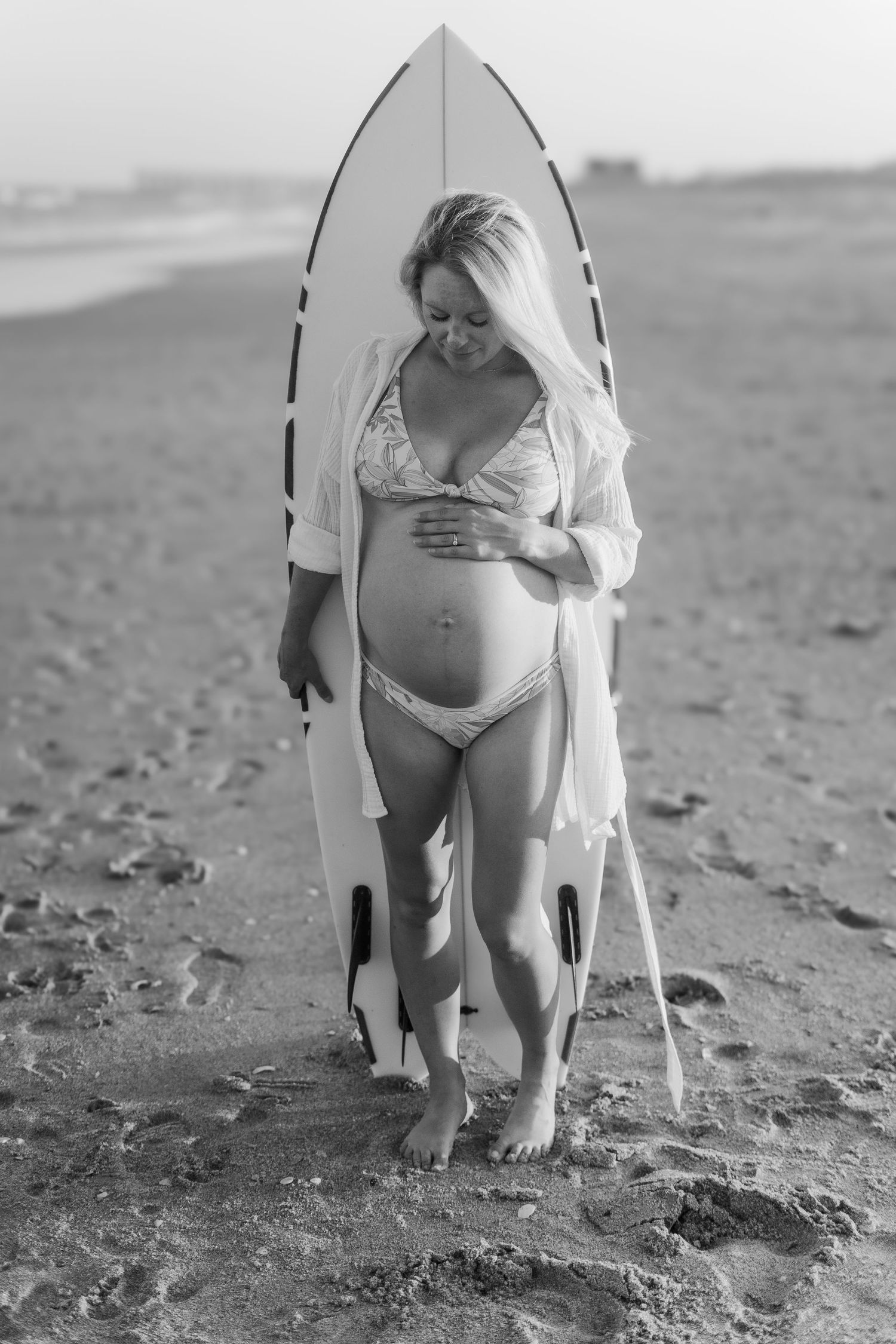 Black and white portrait of pregnant woman with surfboard on beach.