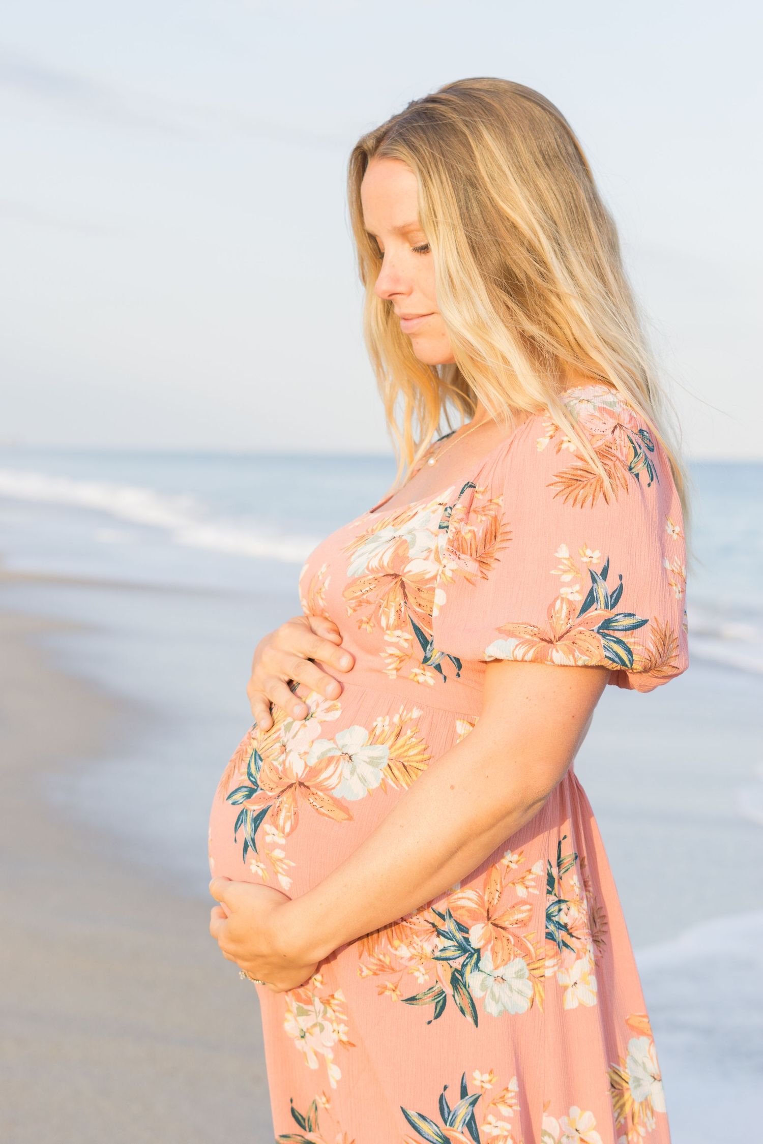 Expecting mother in floral dress cradles baby bump while standing on beach at sunset.