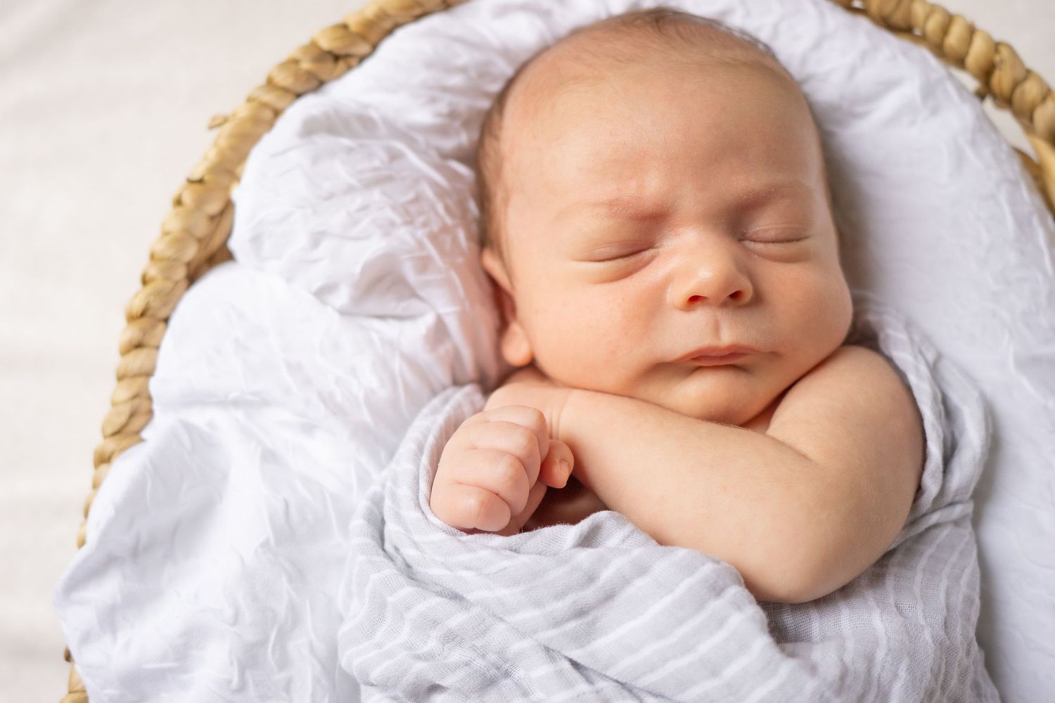 Newborn resting in woven basket with white blanket and peaceful expression.