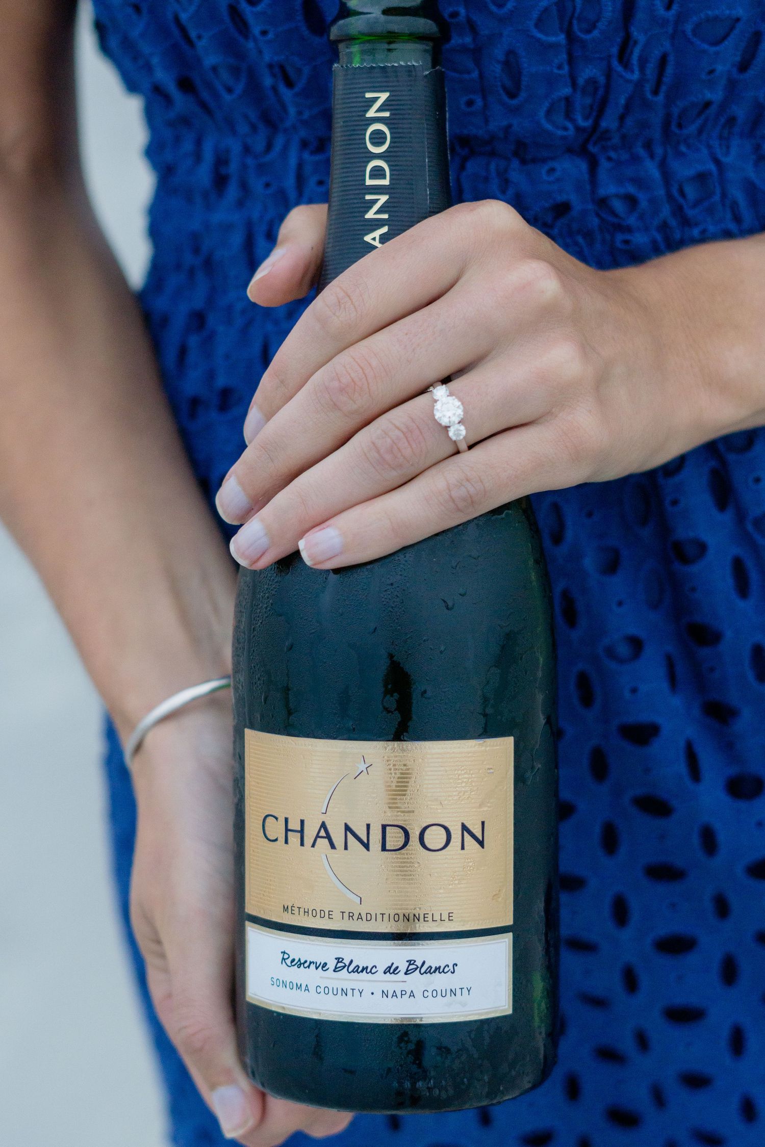 A close-up of hands holding a Chandon champagne bottle showing an engagement ring.