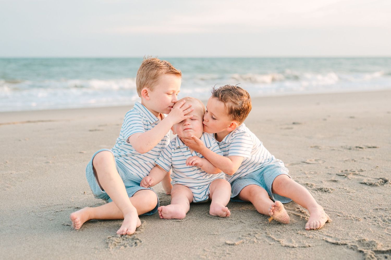 Three siblings in striped shirts sharing a sweet moment together on the beach.