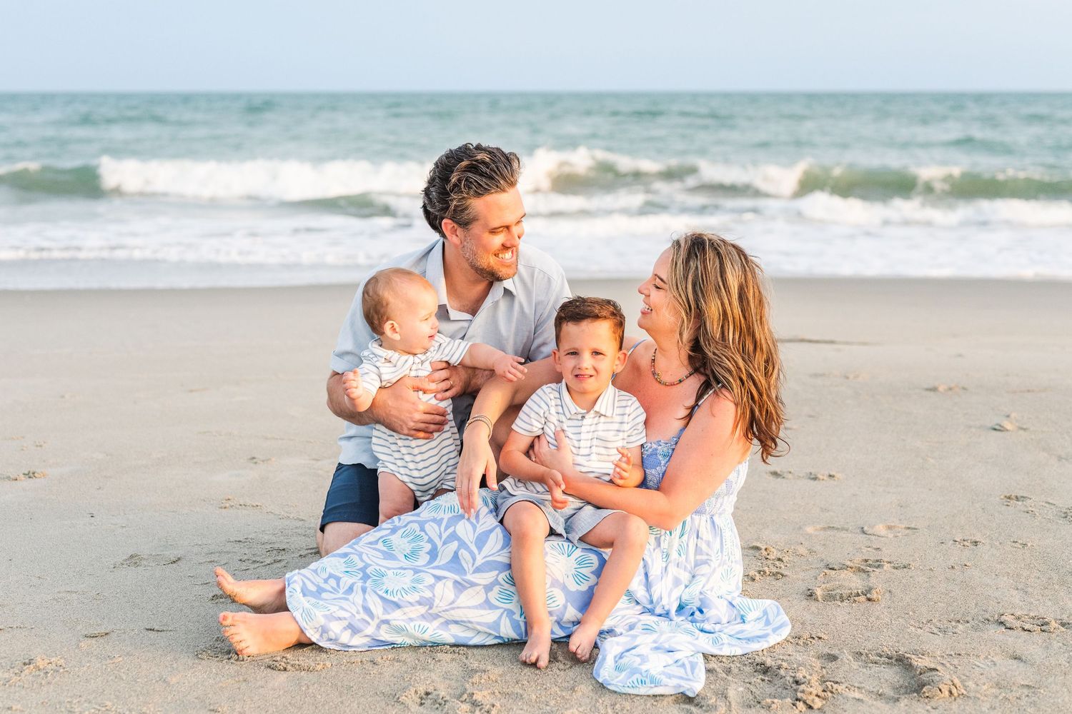 A family group sits together on a sunny beach wearing coordinating blue and white outfits by the ocean waves.