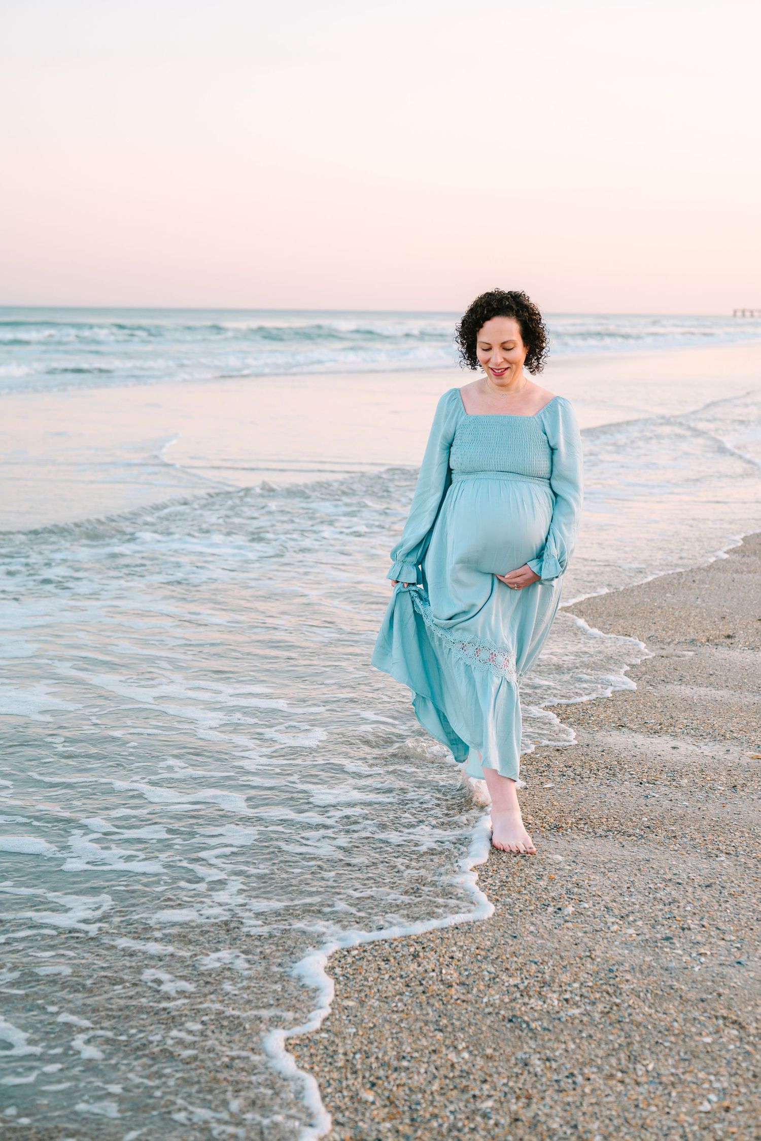 Expectant parent in flowing blue dress walking along beach shoreline at sunset while waves gently lap at the sand.