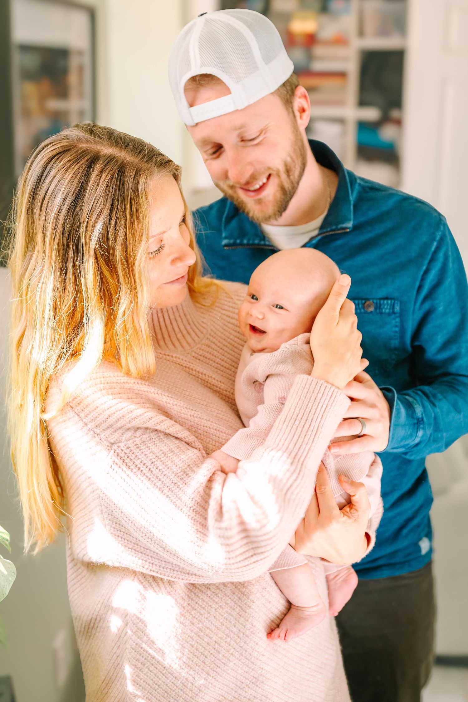 A young couple in casual attire share a sweet moment with their infant baby in a sunlit room.