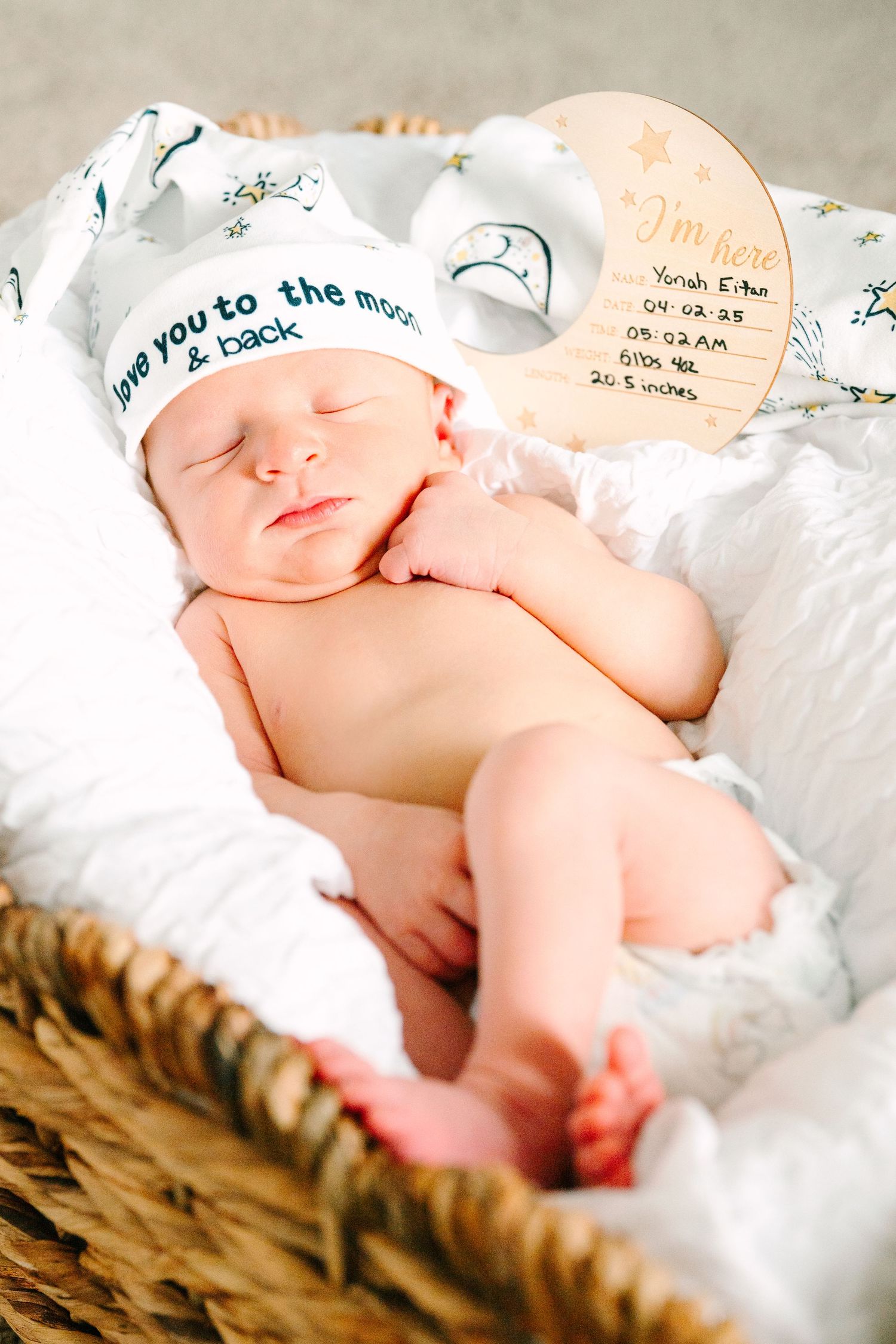 Sleeping newborn wearing hat with 'Love you to the moon and back' text lies in wicker basket on white blanket.