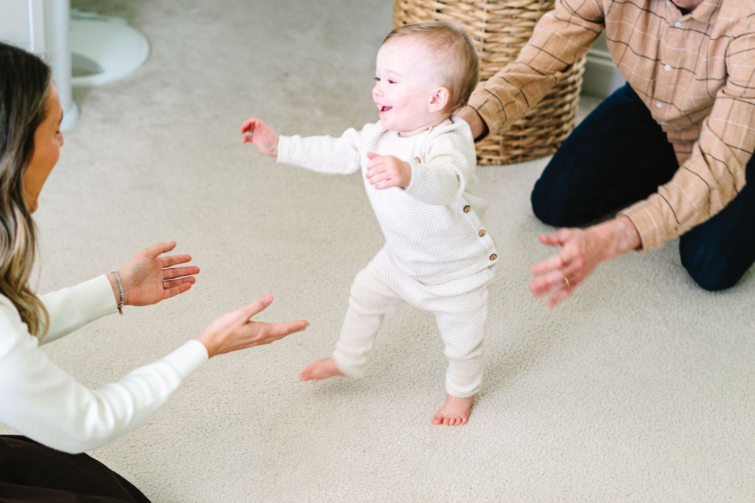 Baby taking first steps in white outfit while parents encourage movement.