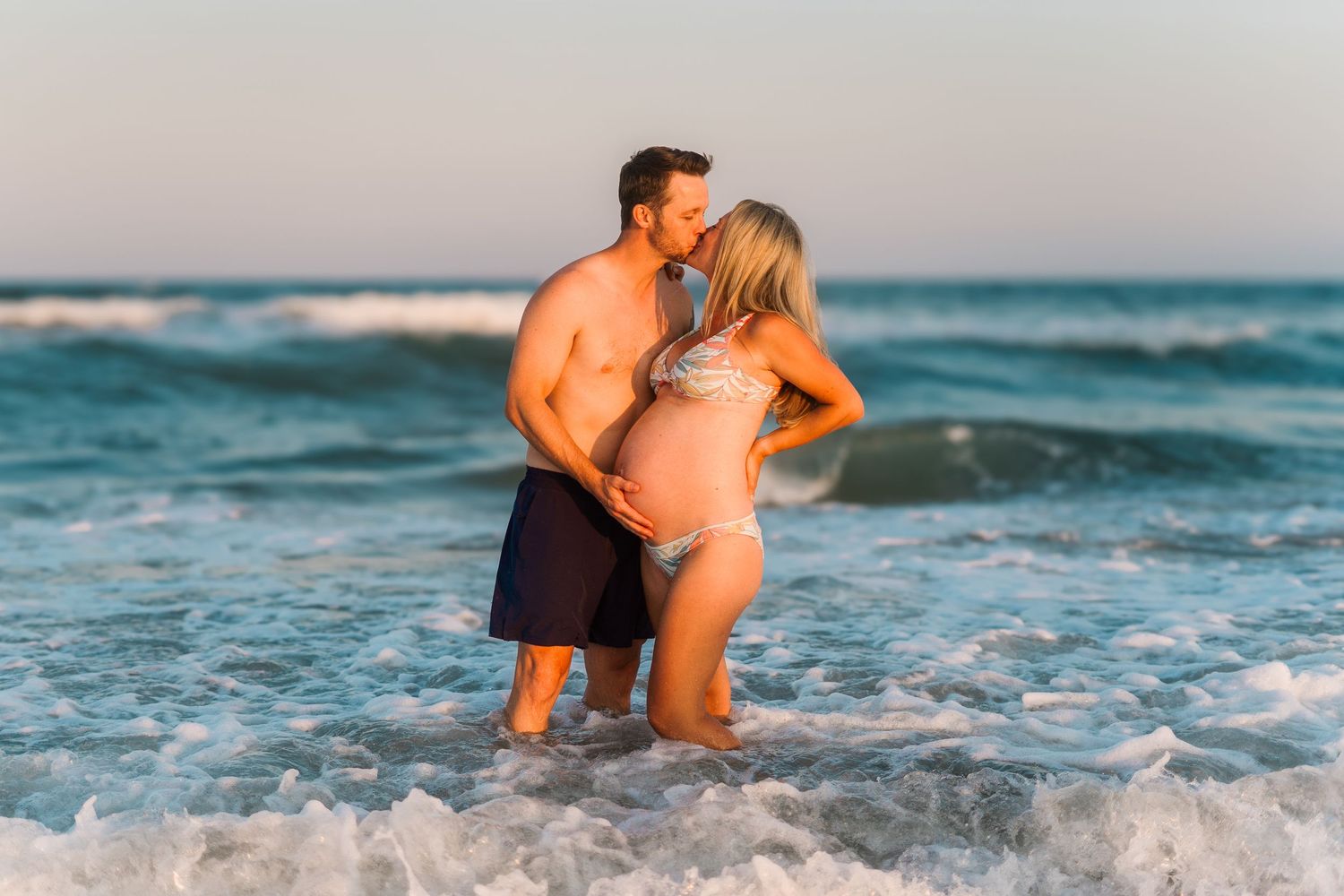 An expecting couple stands in the ocean waves during their beach maternity photoshoot.