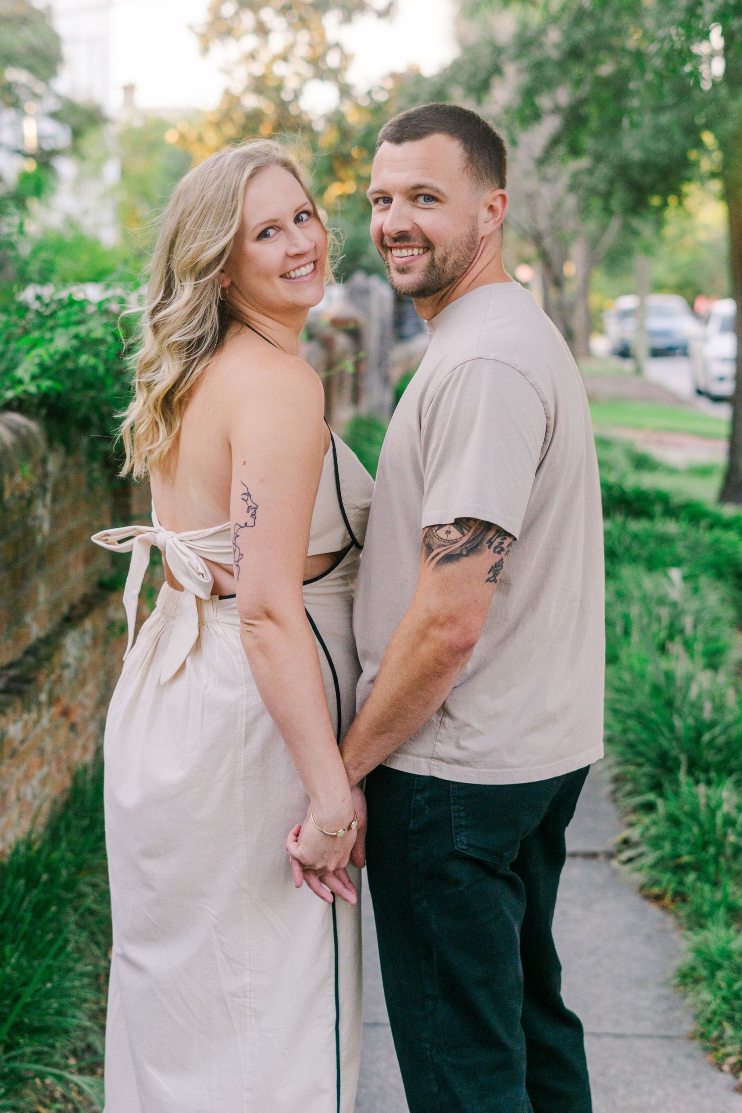 Couple in casual attire poses together on sidewalk surrounded by greenery.