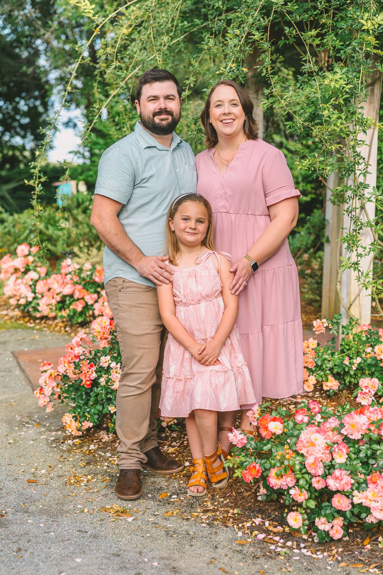 A family poses together on a garden path with blooming coral roses in the background.