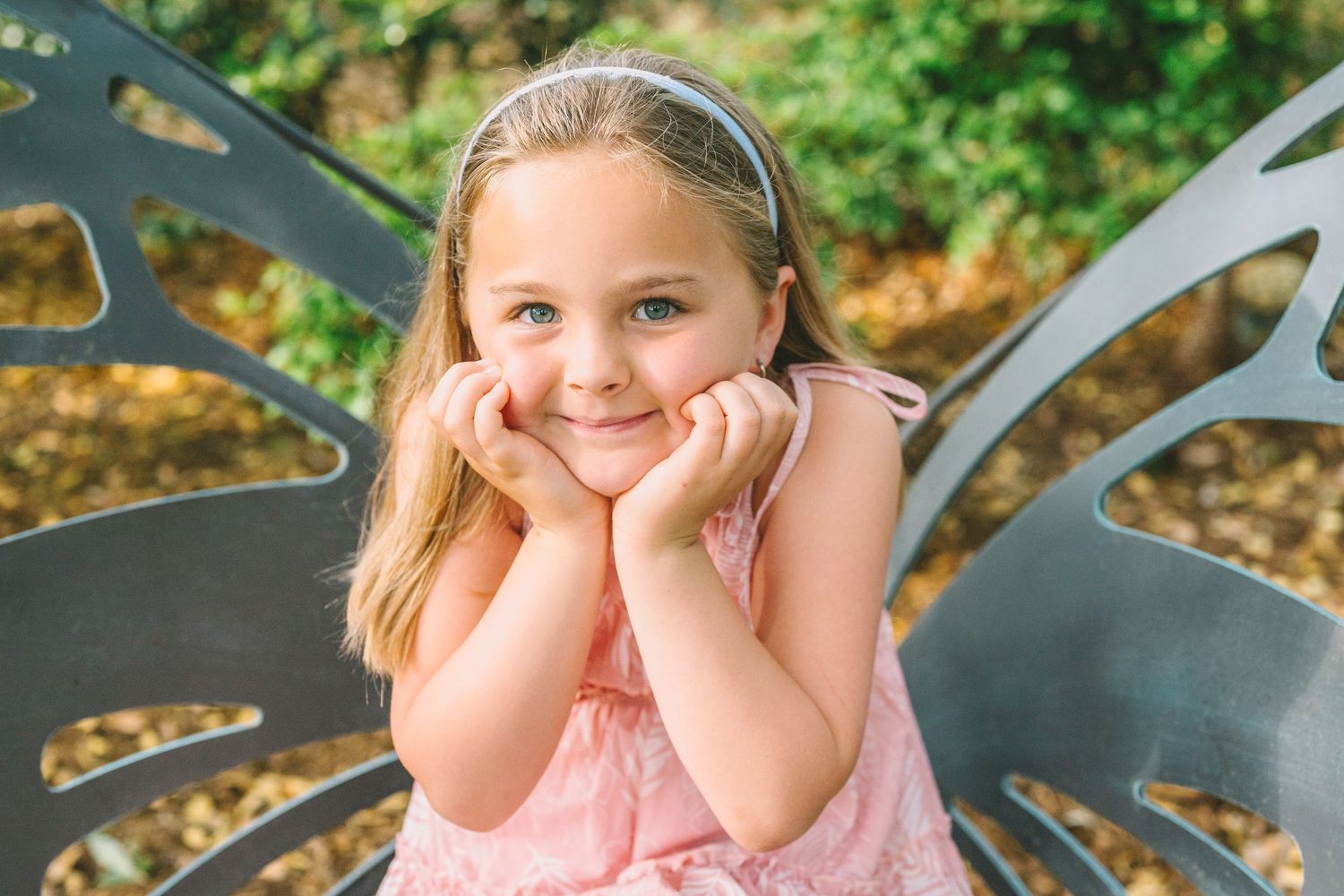 A smiling child in a pink dress sits in a butterfly-shaped chair outdoors with hands propped under their chin.