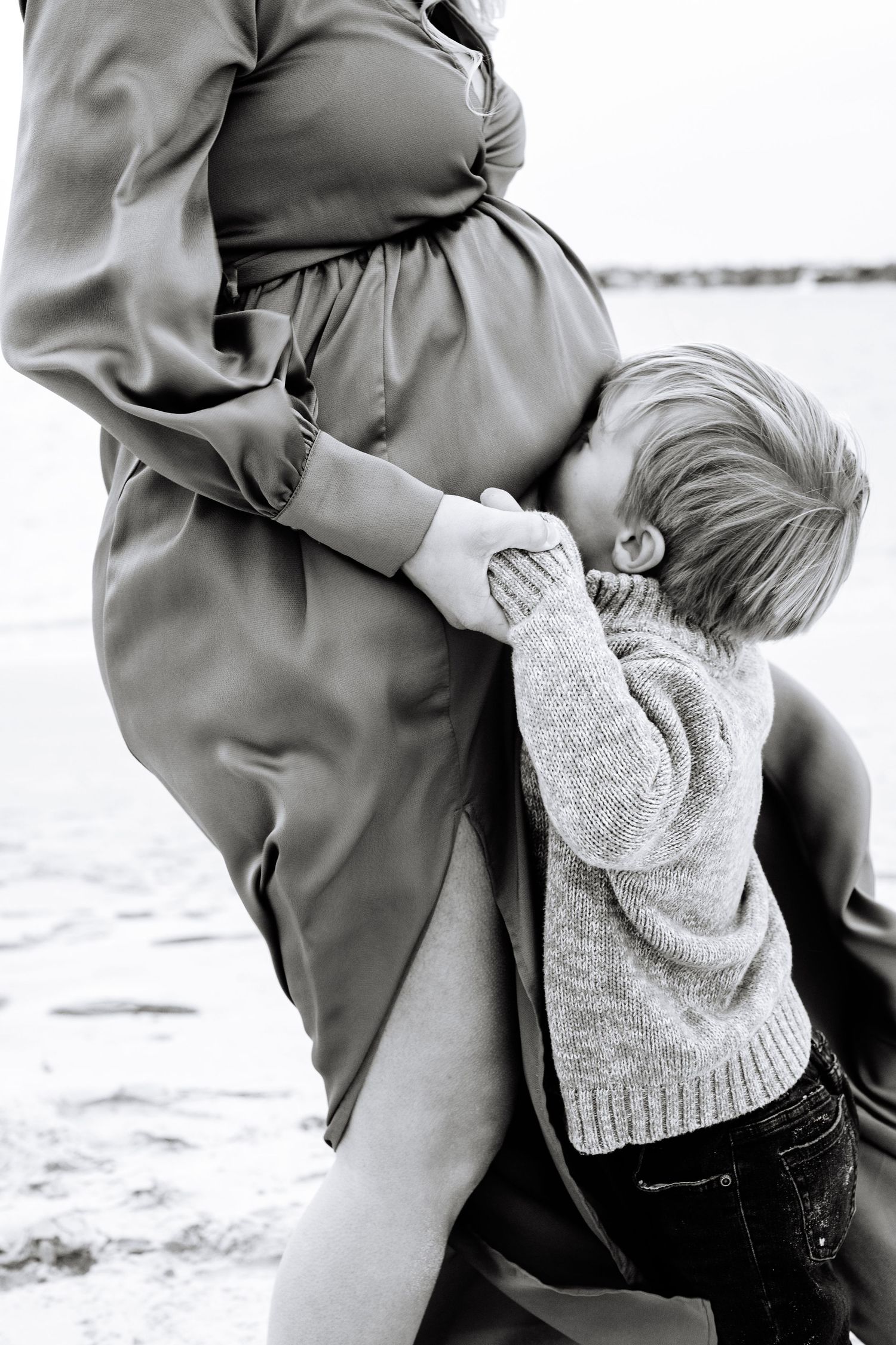 Black and white photo of a child in a knit sweater embracing a pregnant woman's belly outdoors.