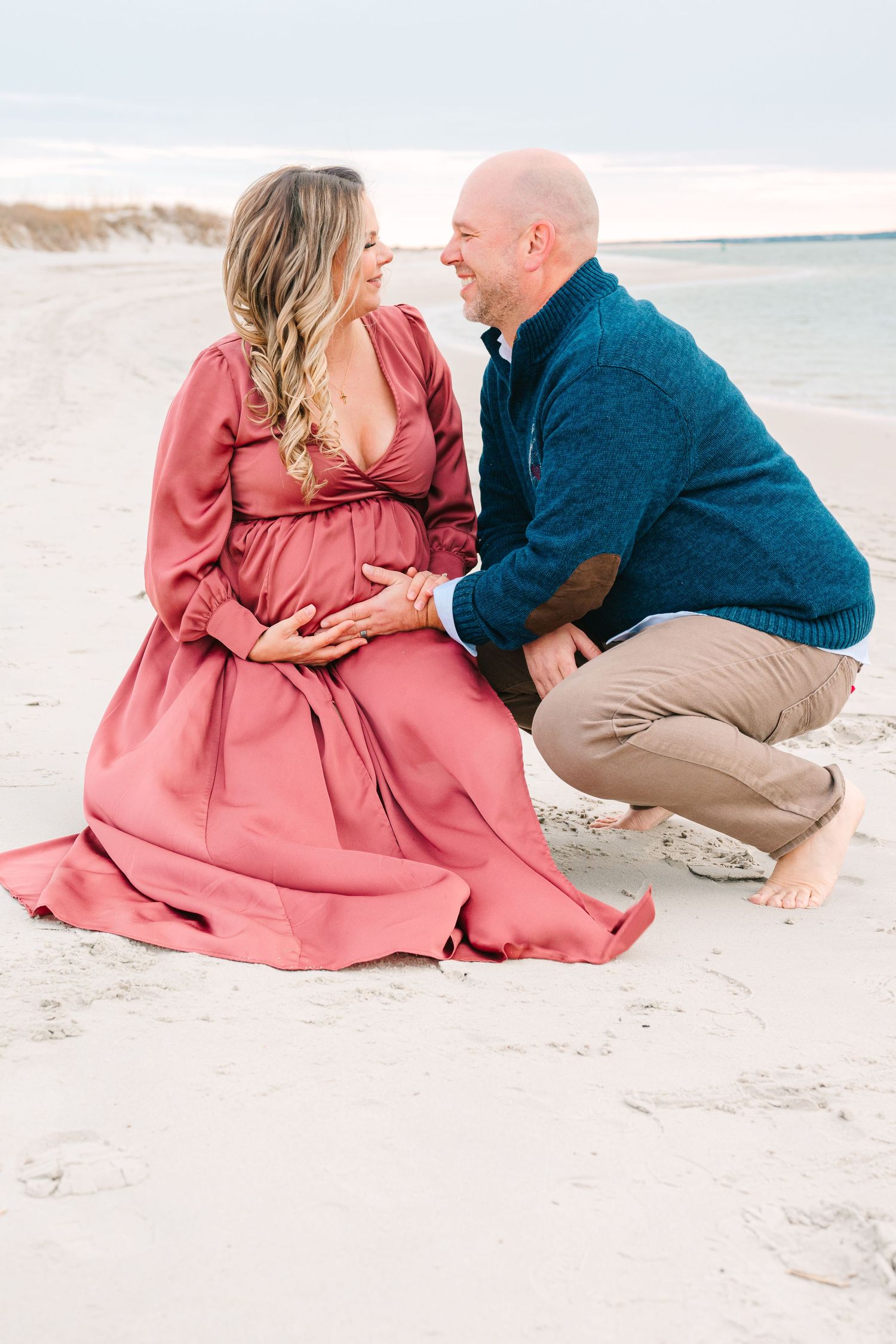 A couple shares an intimate moment on the beach, with her in a flowing mauve dress and him in a blue sweater.