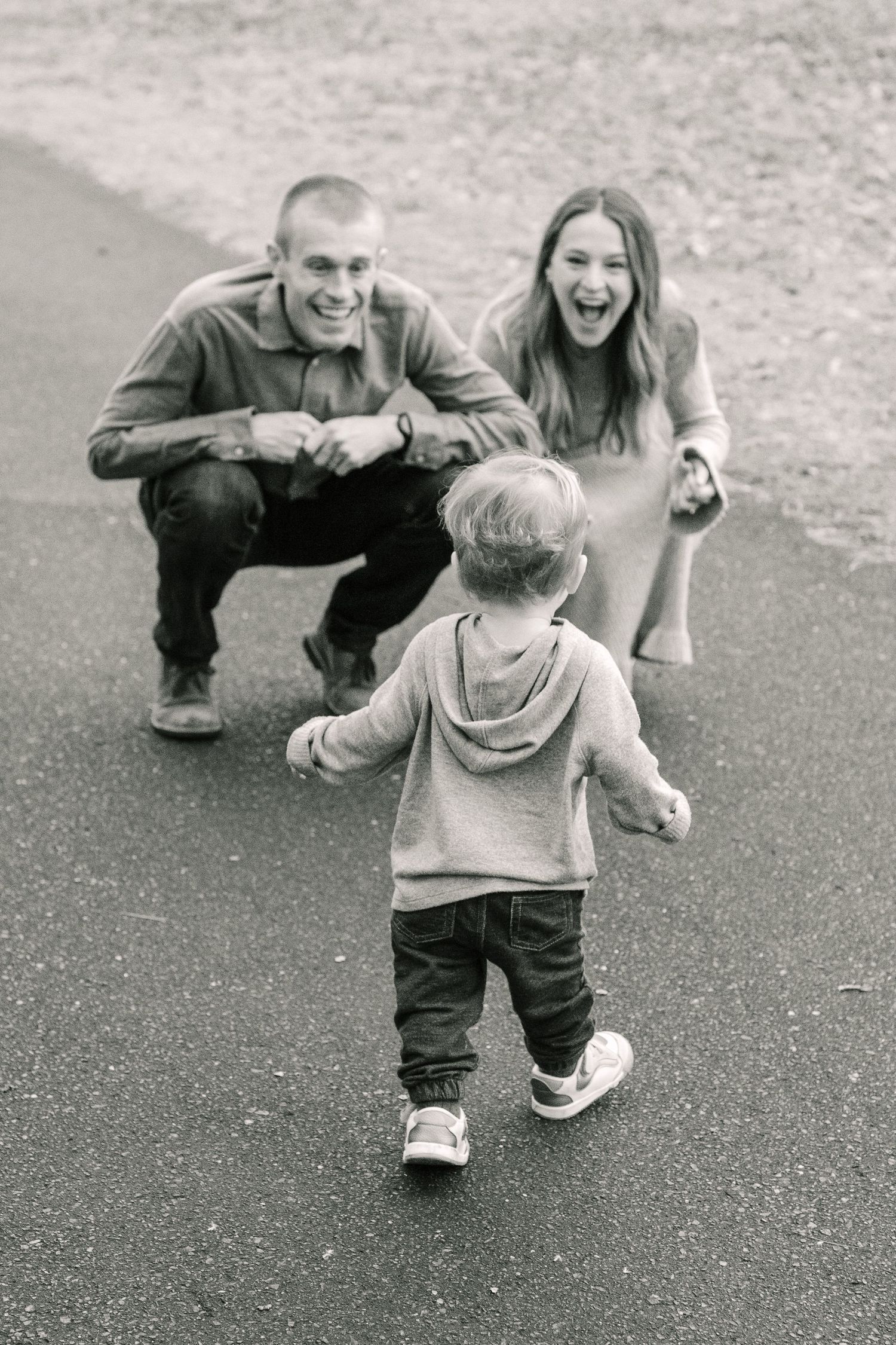 Black and white photo of a toddler in hoodie and sneakers walking towards two adults crouching on pavement.