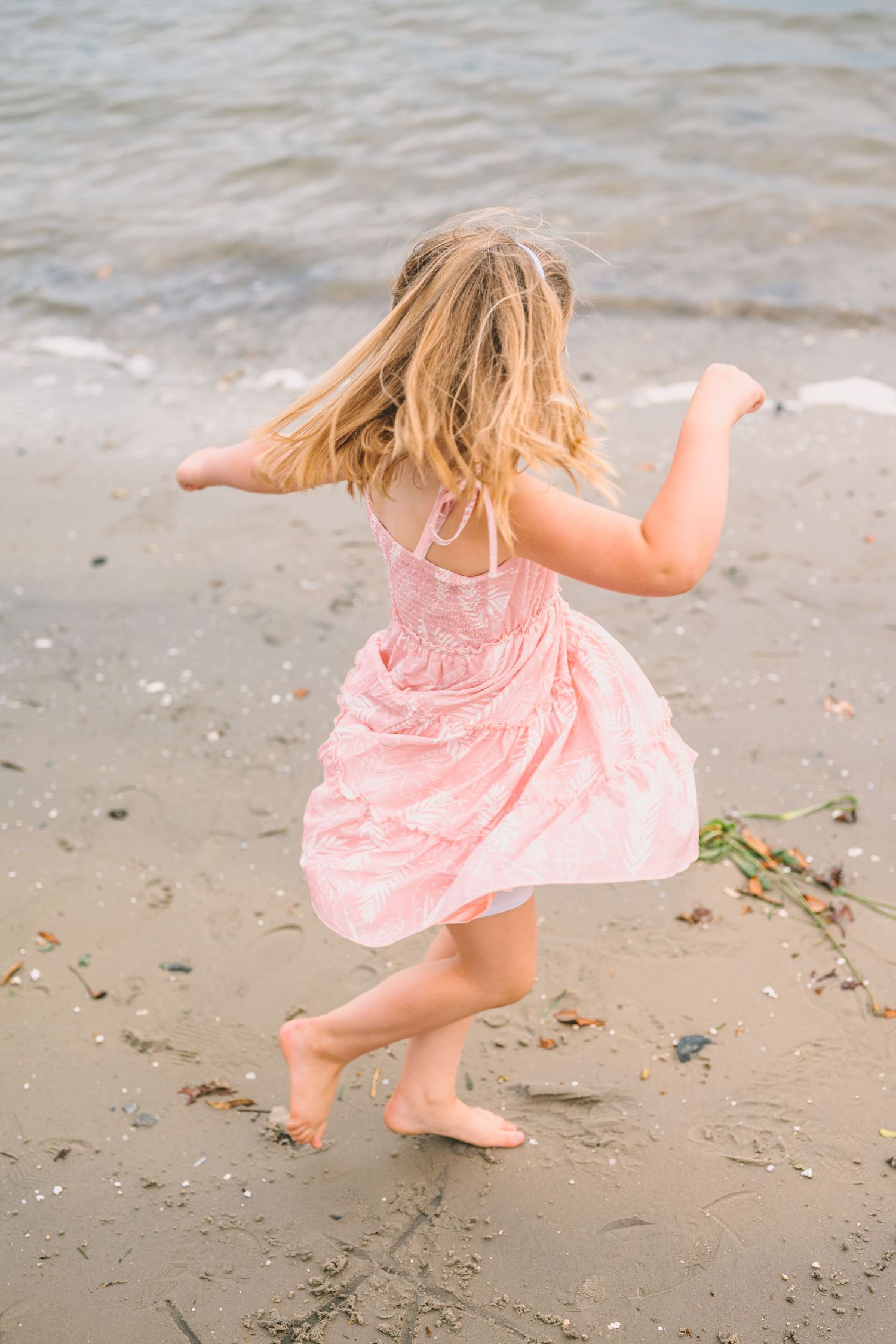 A young child in a pink dress twirls and dances barefoot on the sandy beach.