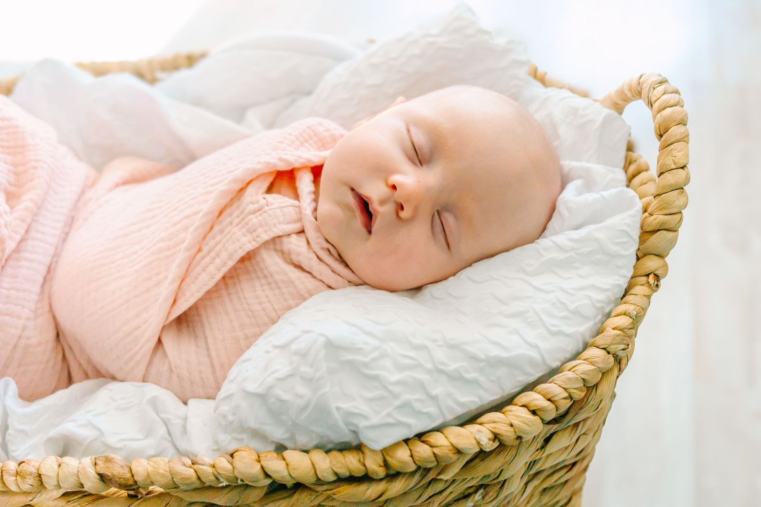 A peaceful sleeping baby in a peach onesie rests in a woven basket on white bedding.