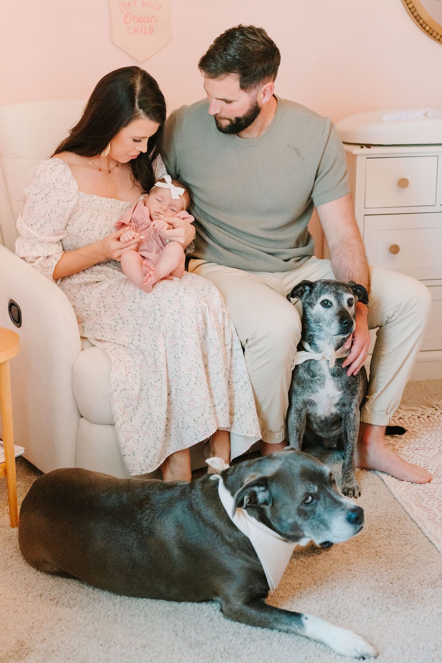 A family sits on a white chair with their baby and two black and white dogs in a pink nursery.