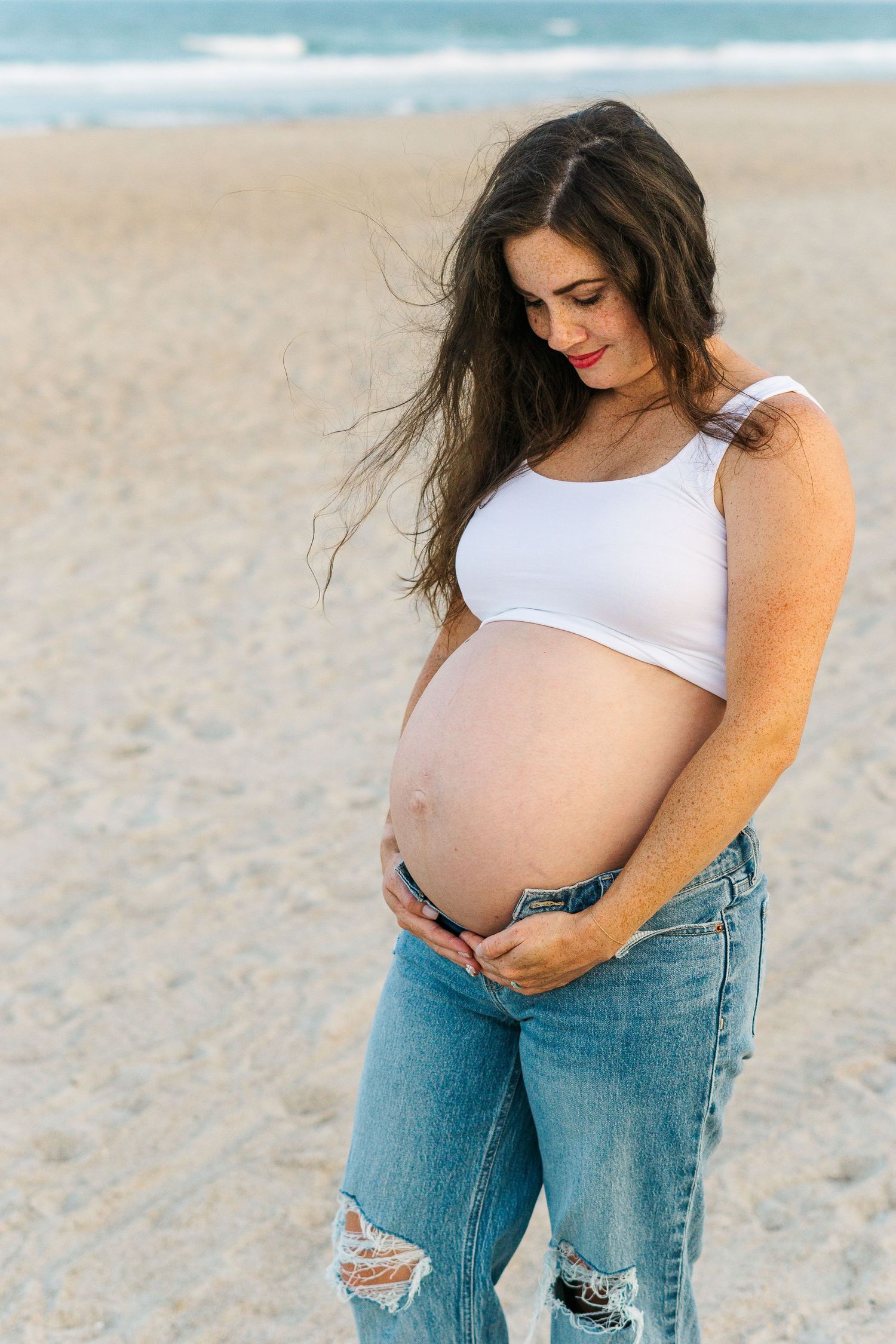 Expecting mother in white crop top and ripped jeans poses on beach with wind-swept hair.
