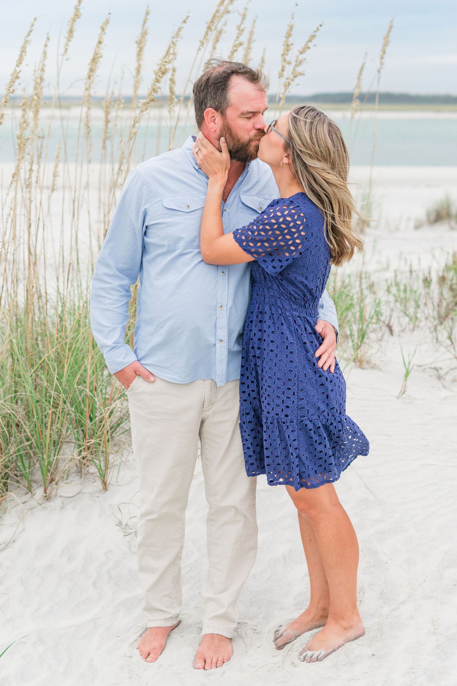 A couple embraces on a sandy beach with sea oats swaying in the foreground.