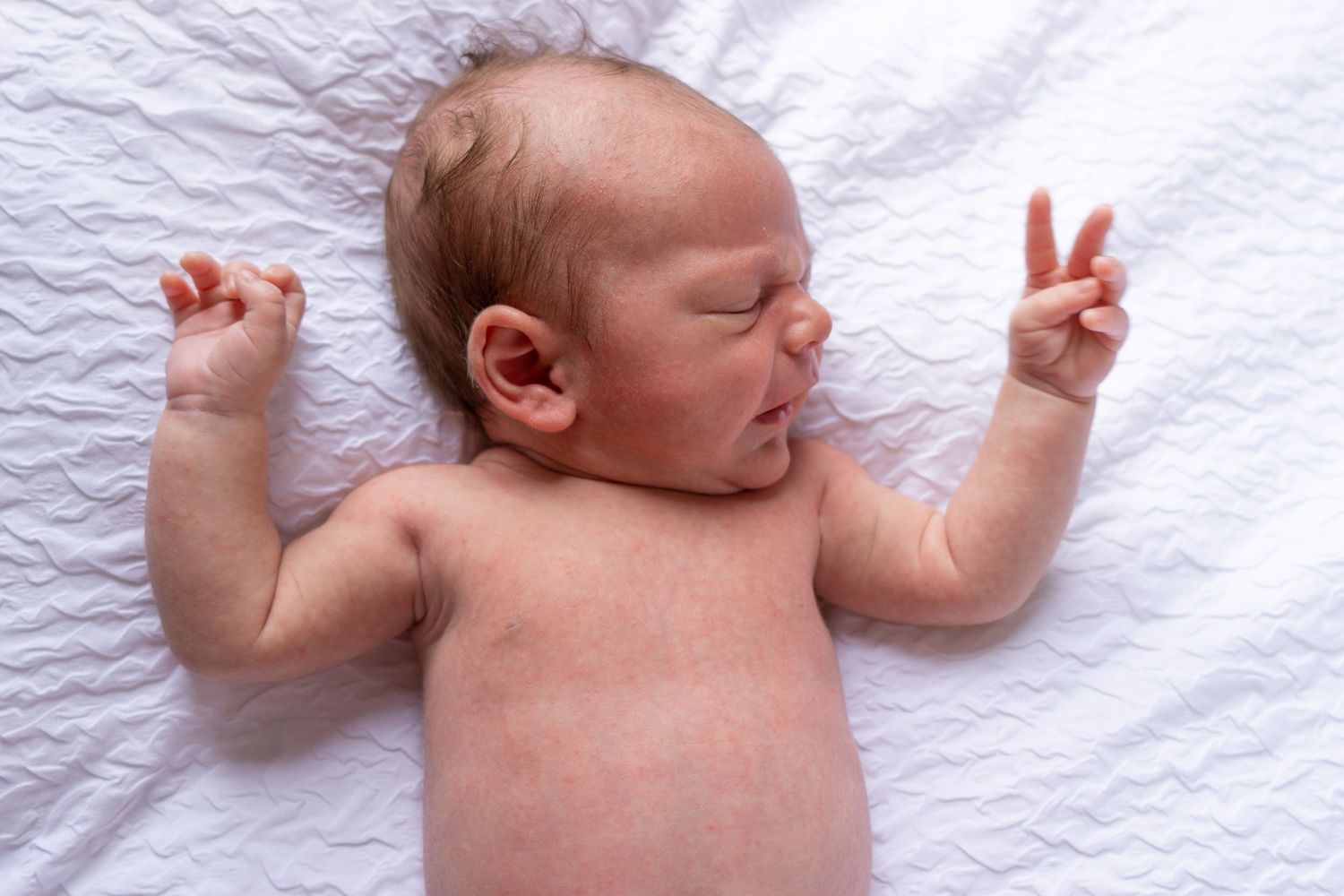Peaceful newborn sleeping on white bedding with arms raised overhead.
