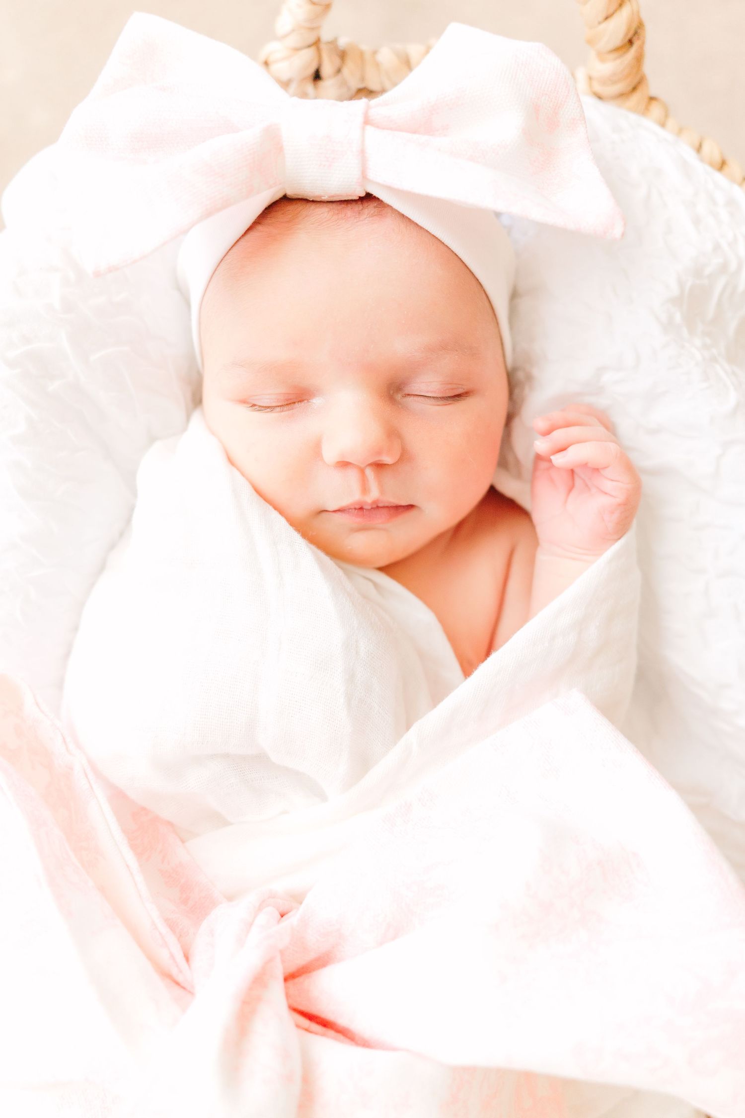 A newborn resting in a woven basket with white wrappings and pink bow accessories.