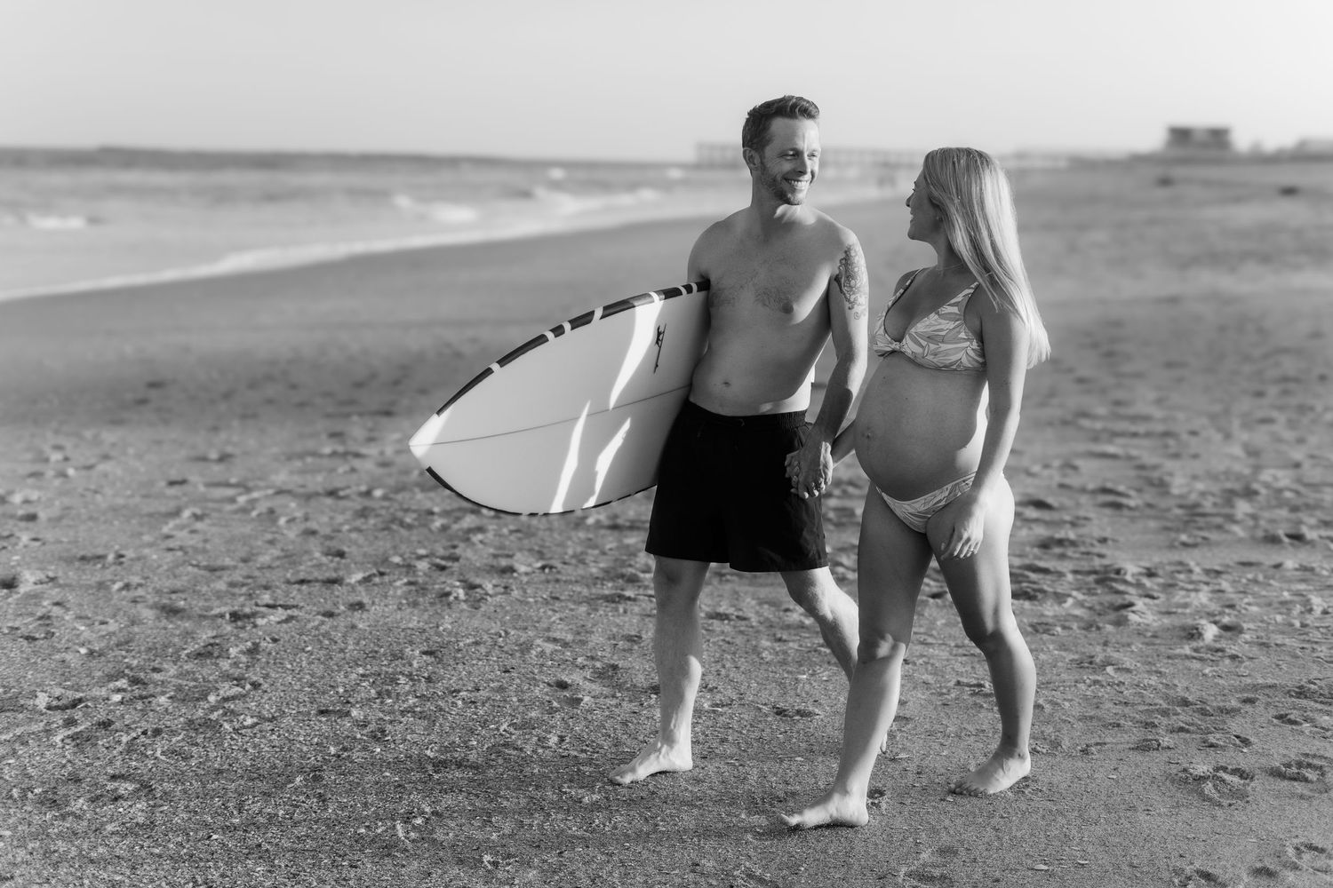 Black and white photo of couple standing with surfboard on sandy beach.
