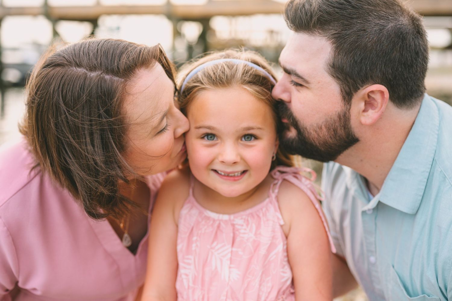 A sweet family moment of parents giving kisses to their child wearing a pink dress.