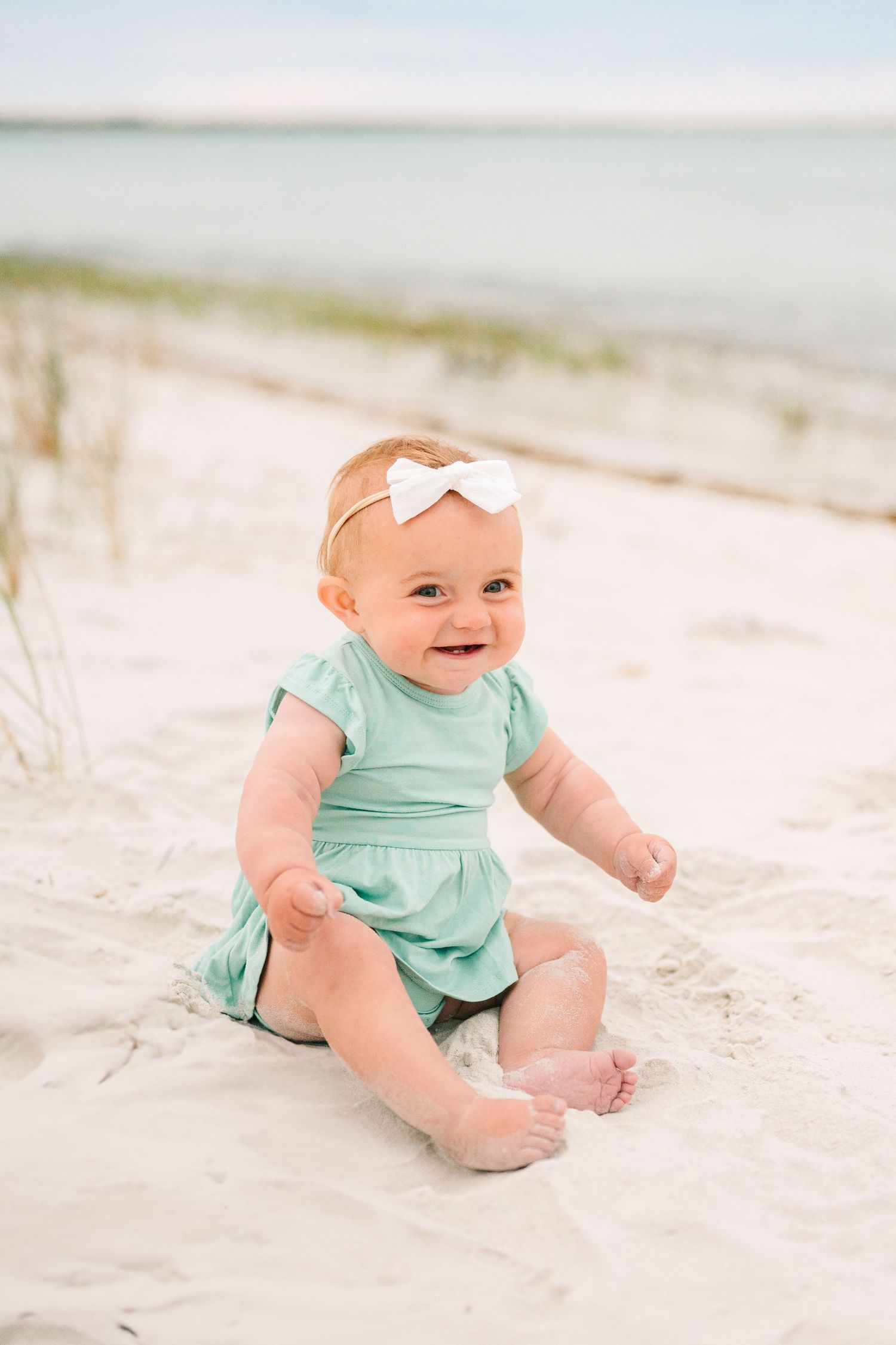 Baby wearing mint green romper and white bow sits happily on sandy beach with dunes in background.