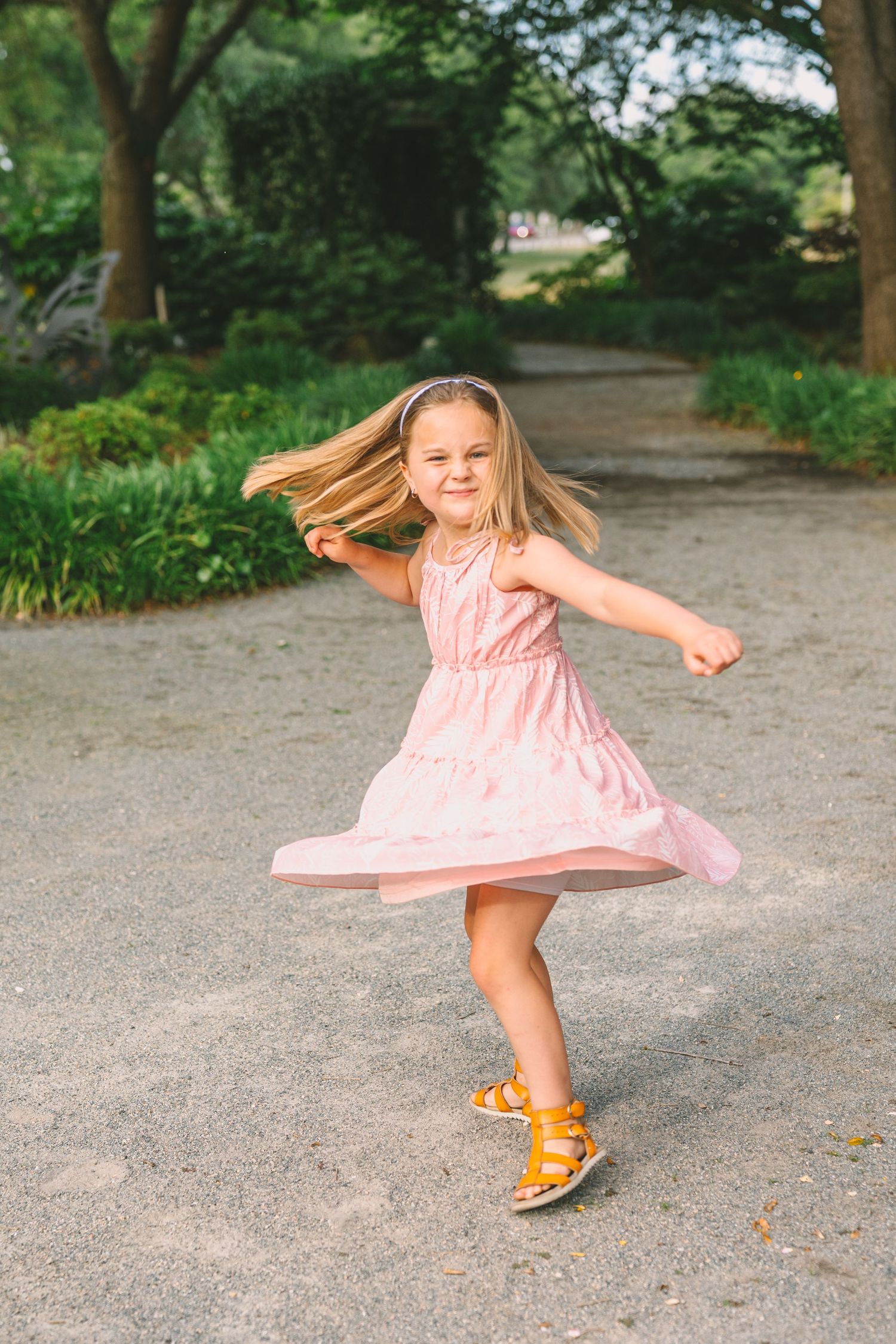 Child in pink dress twirls and dances on a garden path while wearing orange sandals.