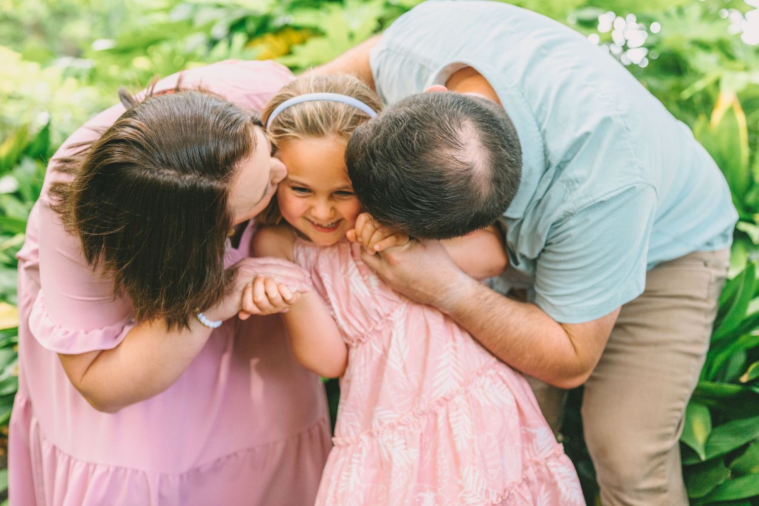 A sweet family moment captured outdoors with affectionate gestures and soft pink clothing against a leafy green background.