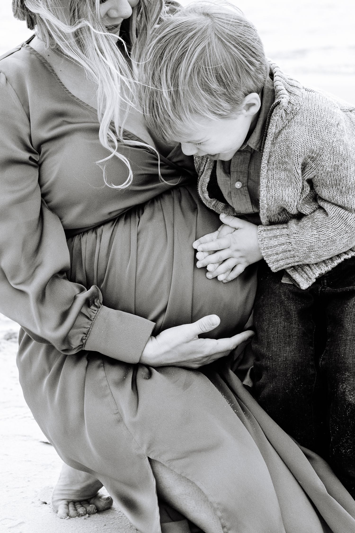 A small child leans tenderly against a pregnant belly in an intimate black and white photograph.