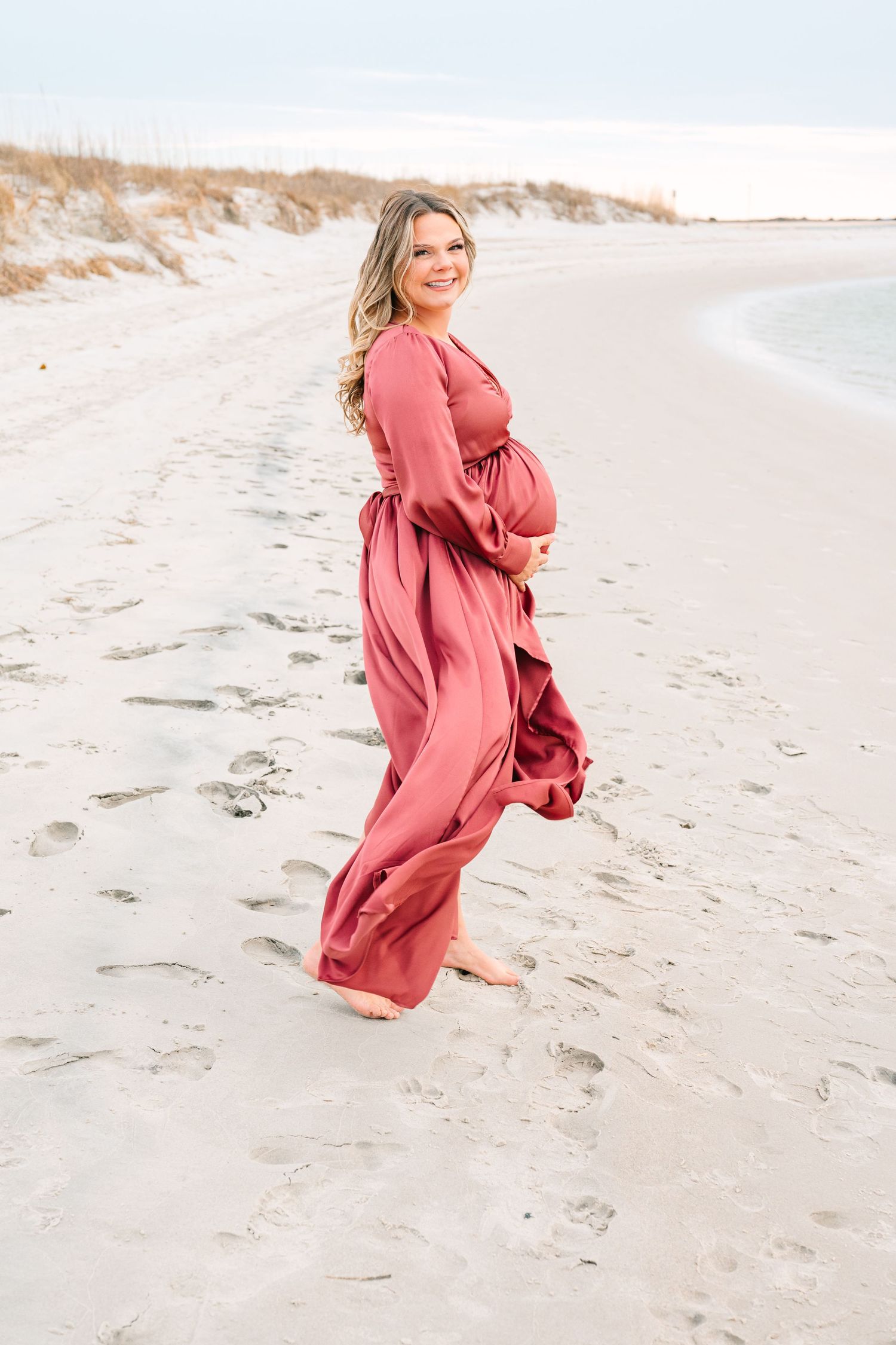 A pregnant figure in a flowing coral maxi dress poses on a sandy beach at sunset.