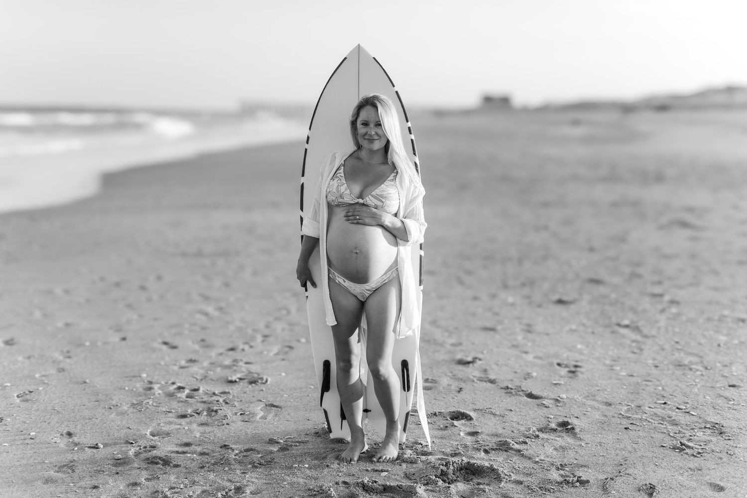 Black and white beach photo of pregnant woman standing with surfboard.