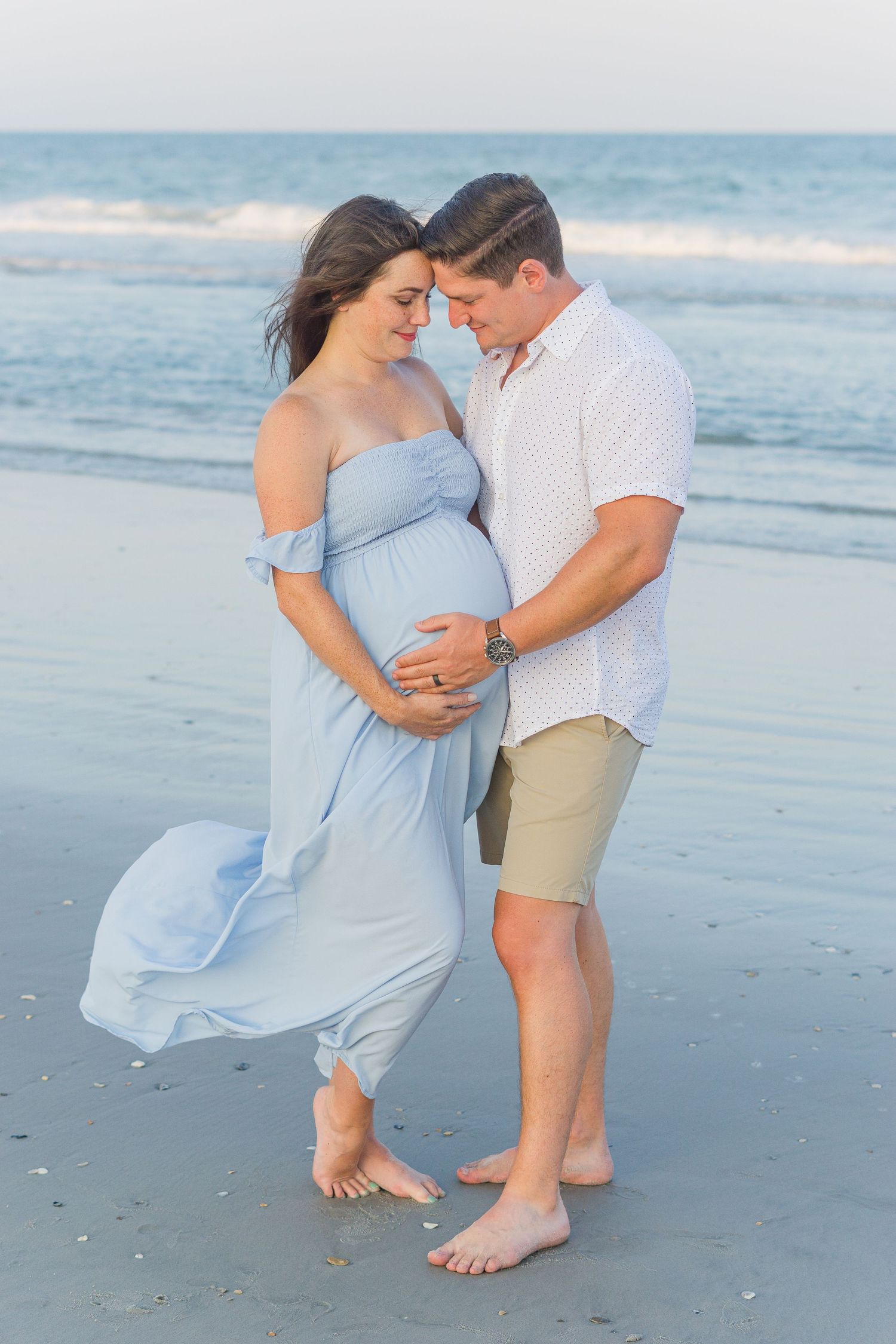 Intimate beachside maternity portrait with flowing light blue dress catching the breeze.