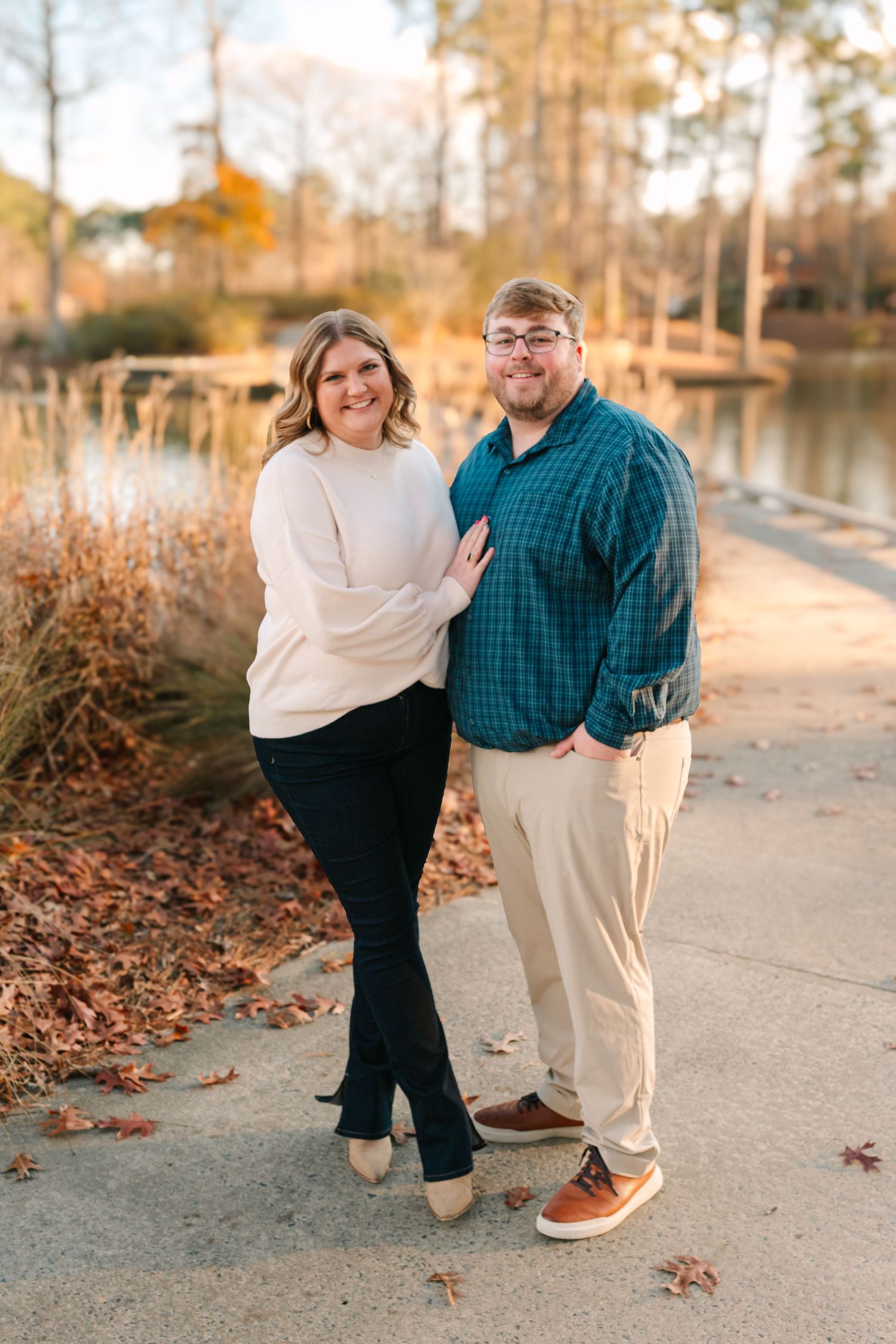 A couple poses together on a path by a lake during autumn sunset.