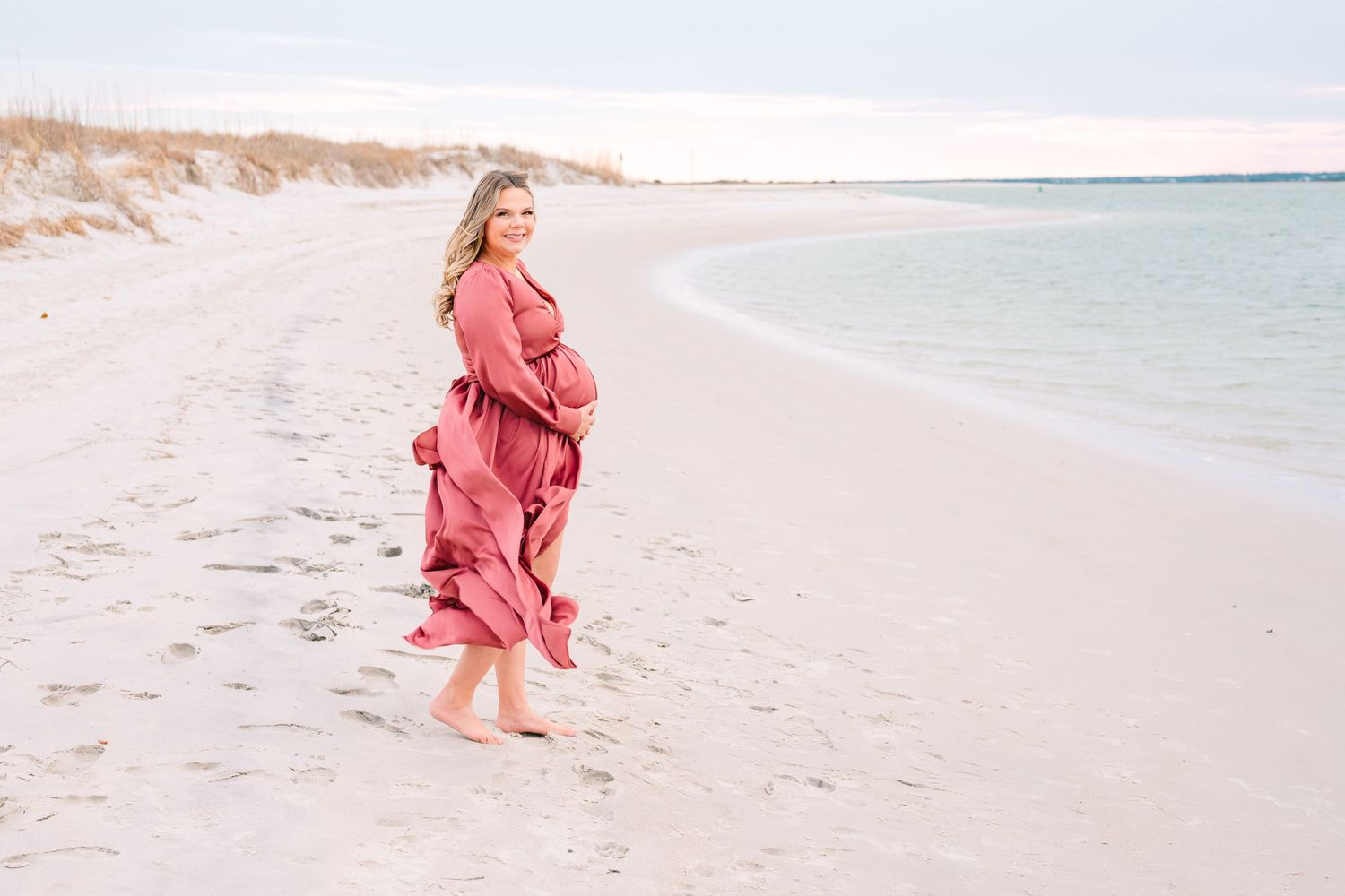 Person in flowing coral dress walking barefoot on sandy beach with ocean waves in background during sunset.