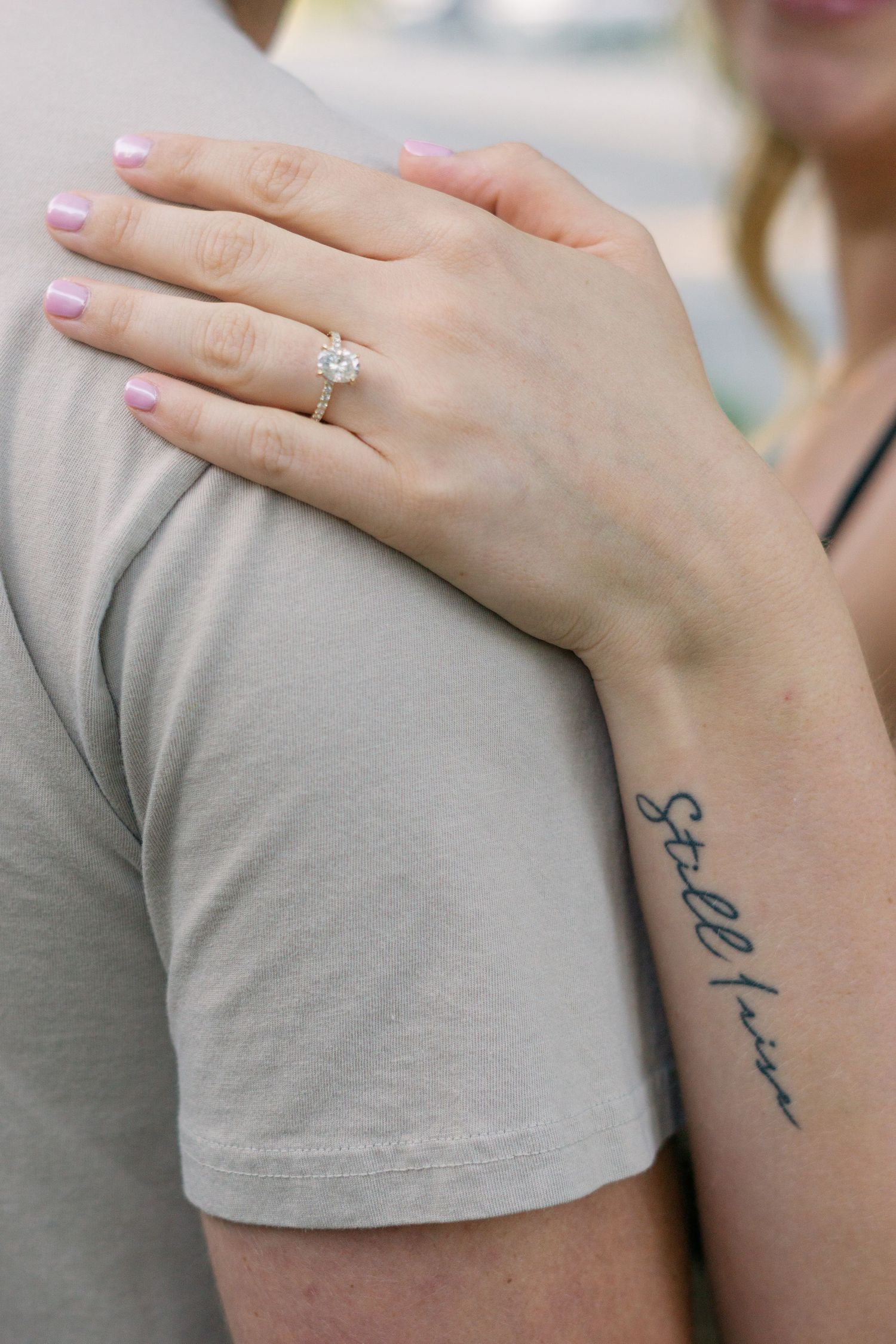 Close-up of engagement ring and script tattoo on arm against grey shirt.