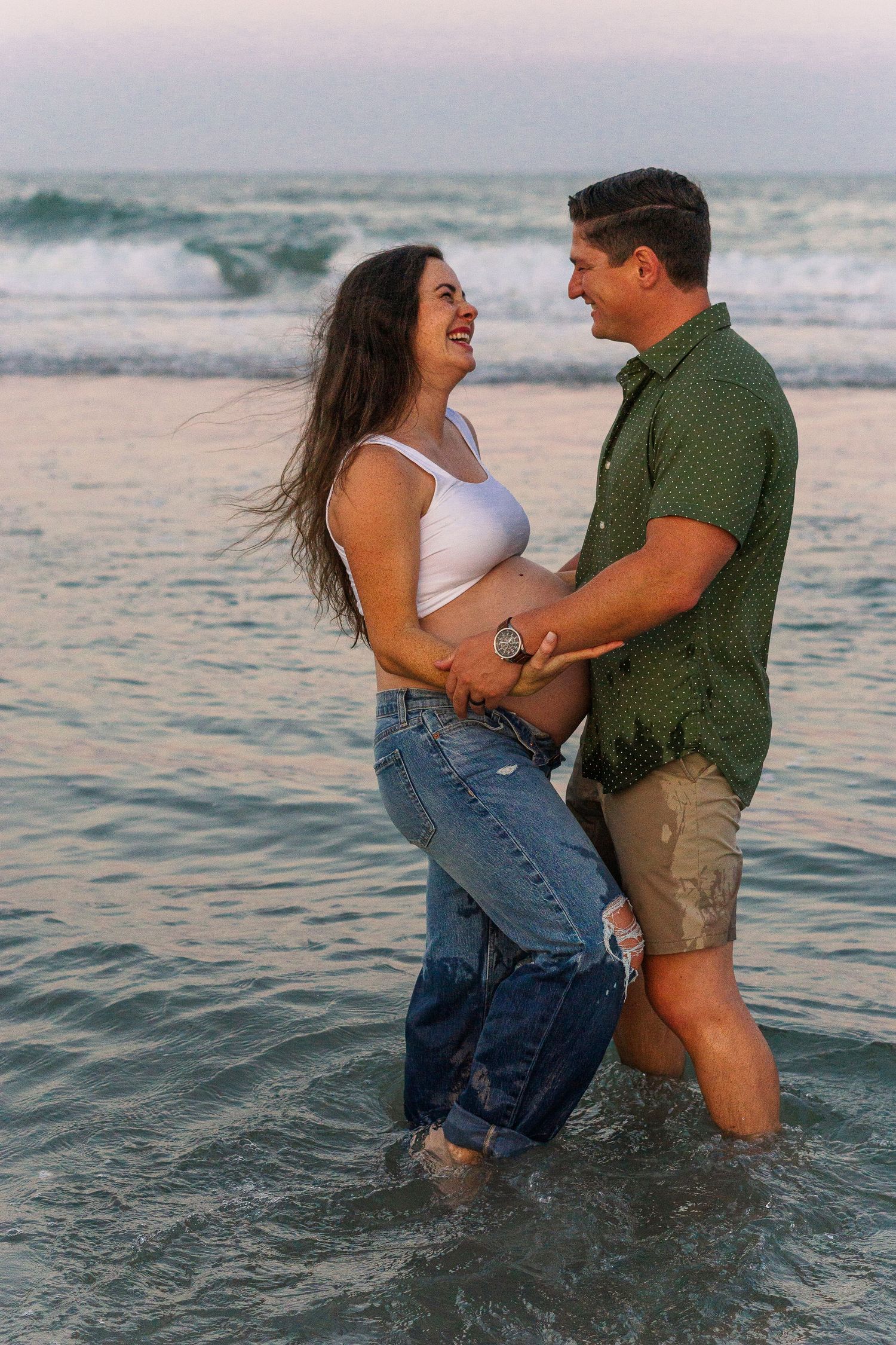 Joyful couple laughing together while standing in shallow ocean water during sunset.