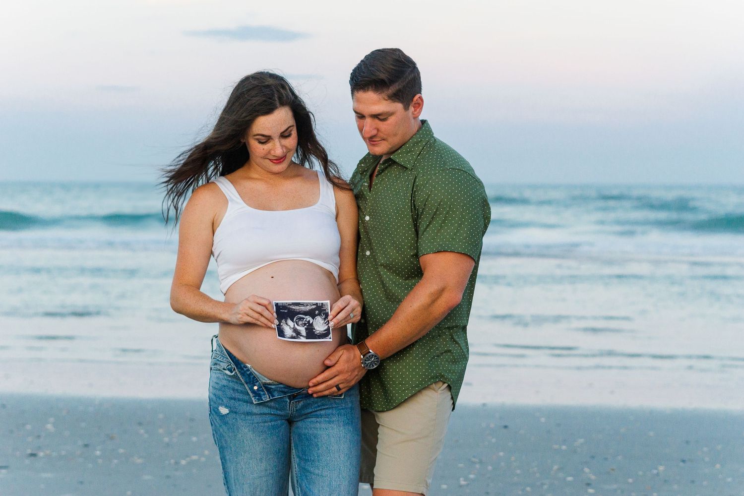 A couple shares a tender moment while holding an ultrasound photo on the beach.