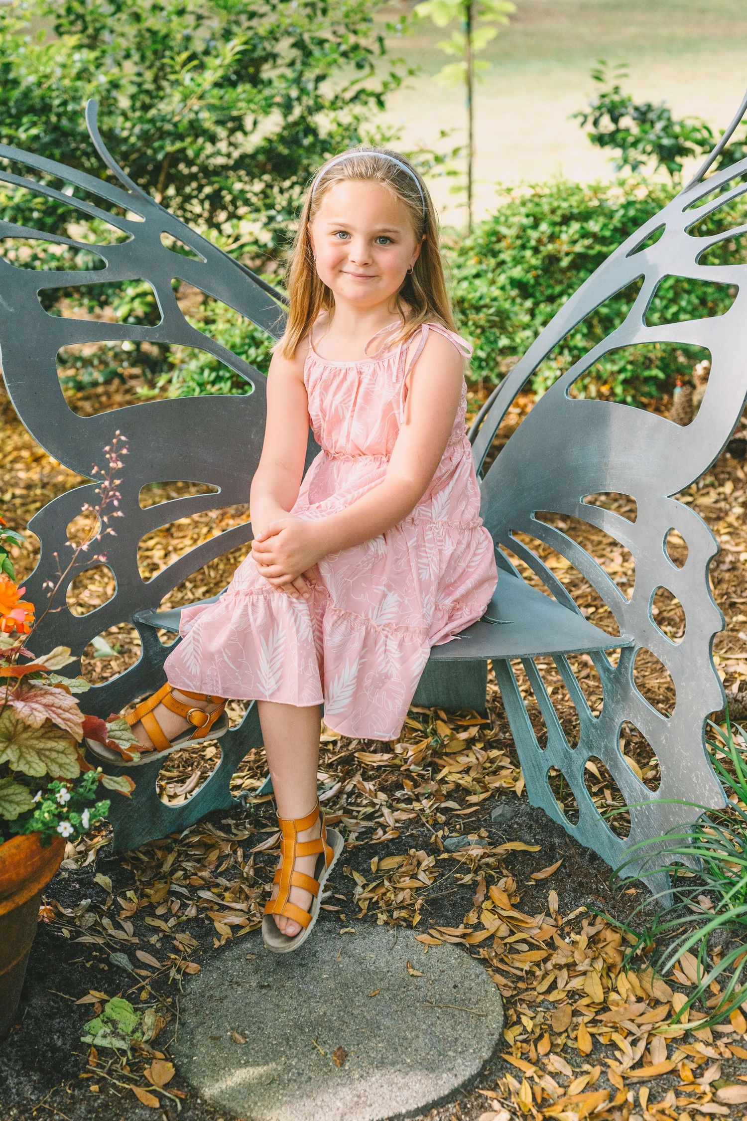 A child in a pink dress sits on a decorative metal butterfly bench in a garden setting.