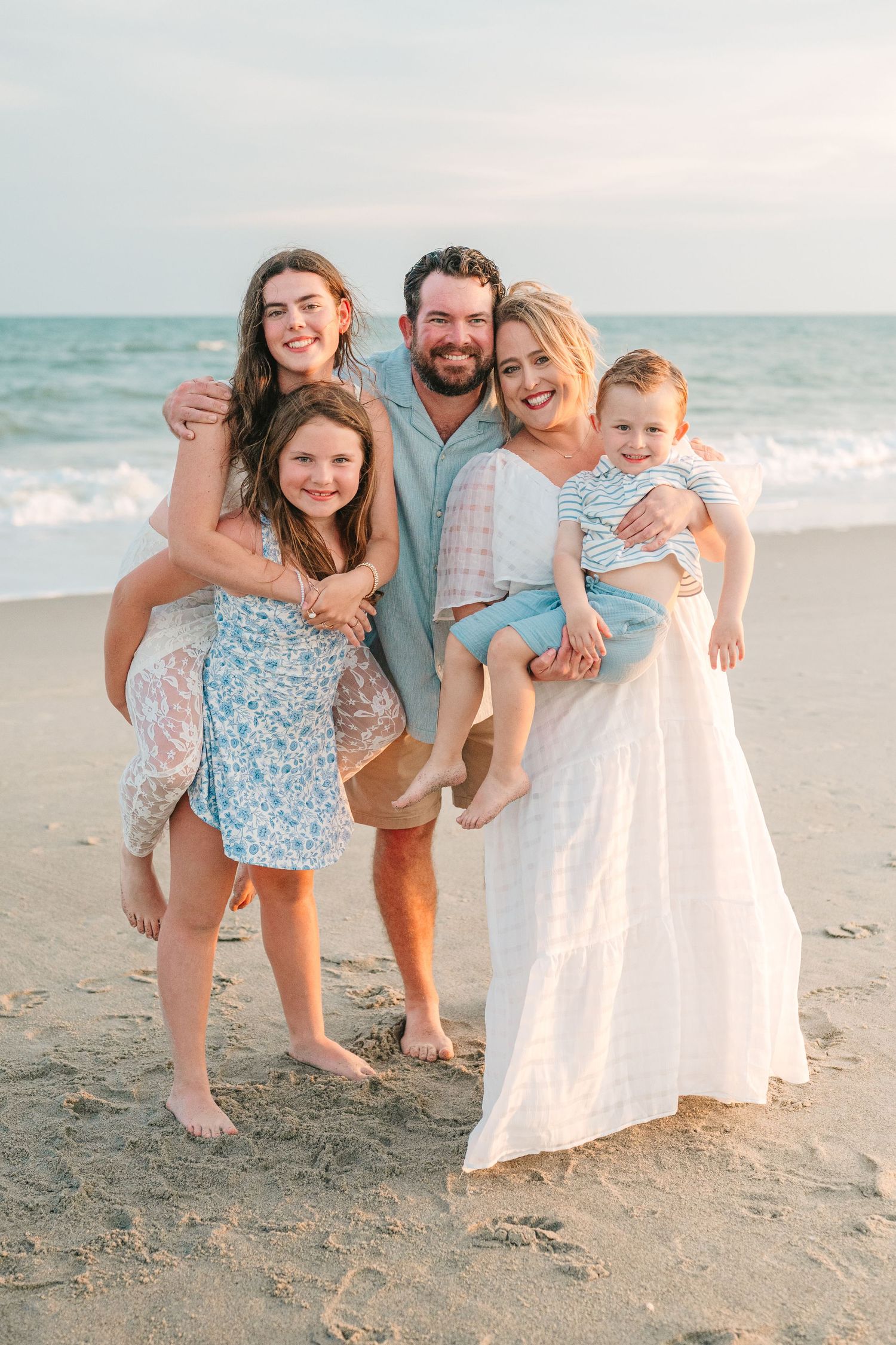Happy family of five embraces on the beach wearing coordinated coastal attire.