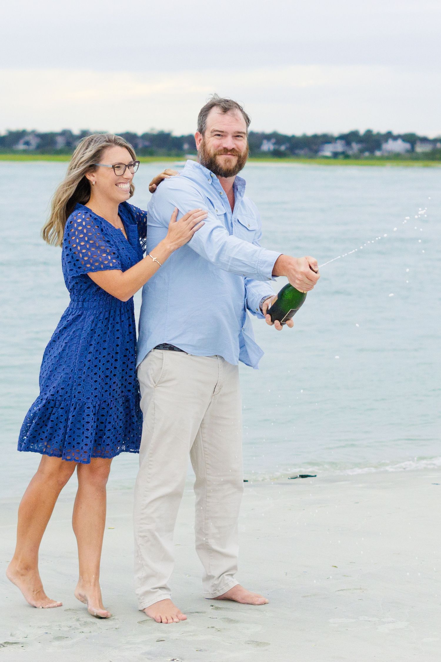 Couple in casual attire celebrates on a sandy beach with champagne as waves roll in behind them.