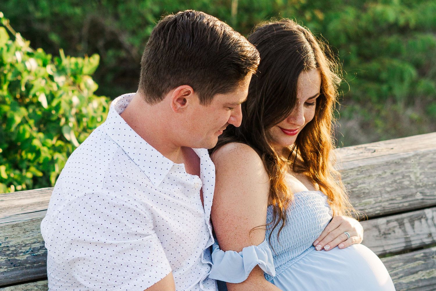 A tender maternity moment shared between expectant parents on a wooden bench outdoors.