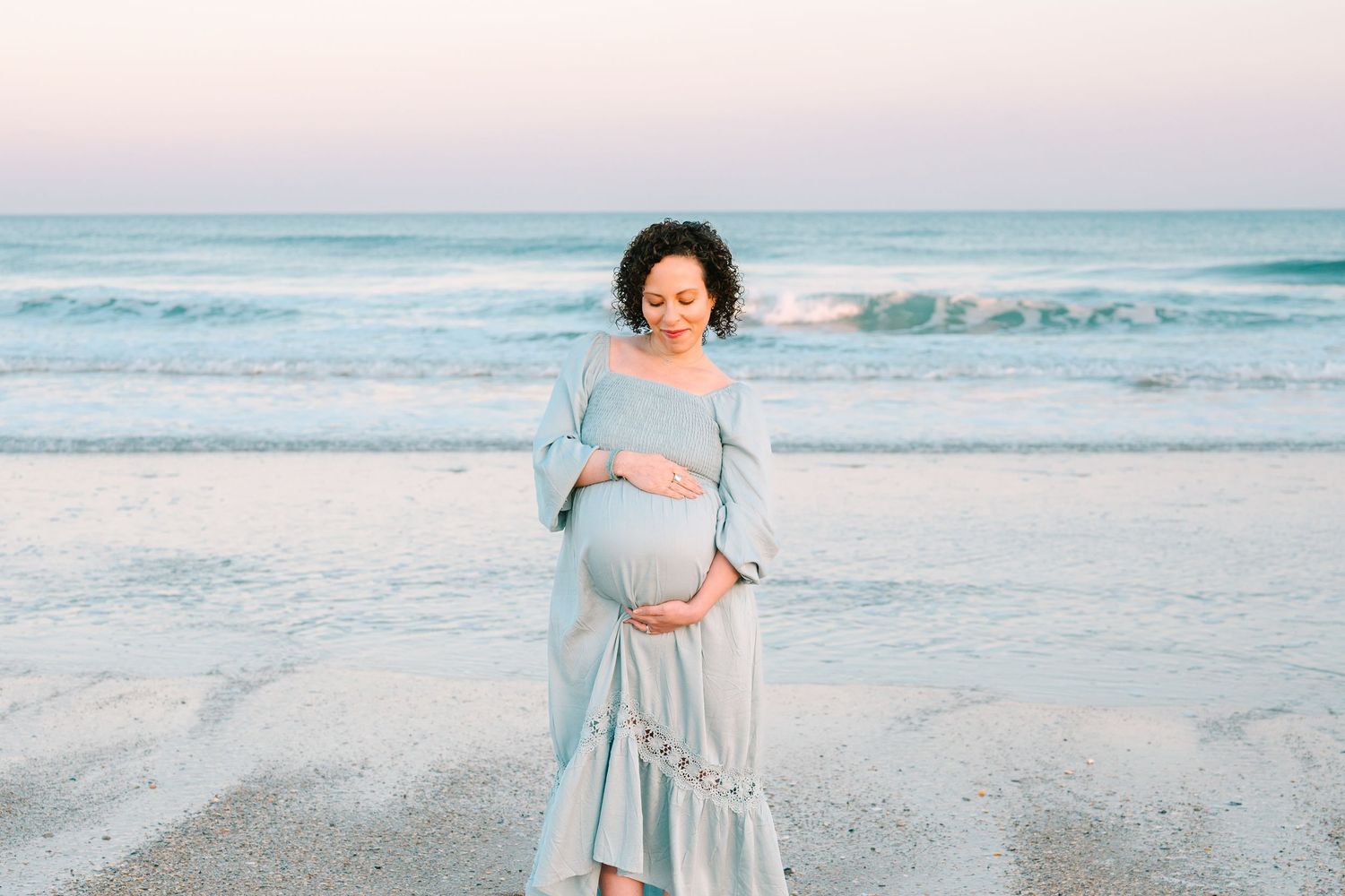 A person in a flowing blue dress stands on a beach at sunset while cradling their pregnant belly.